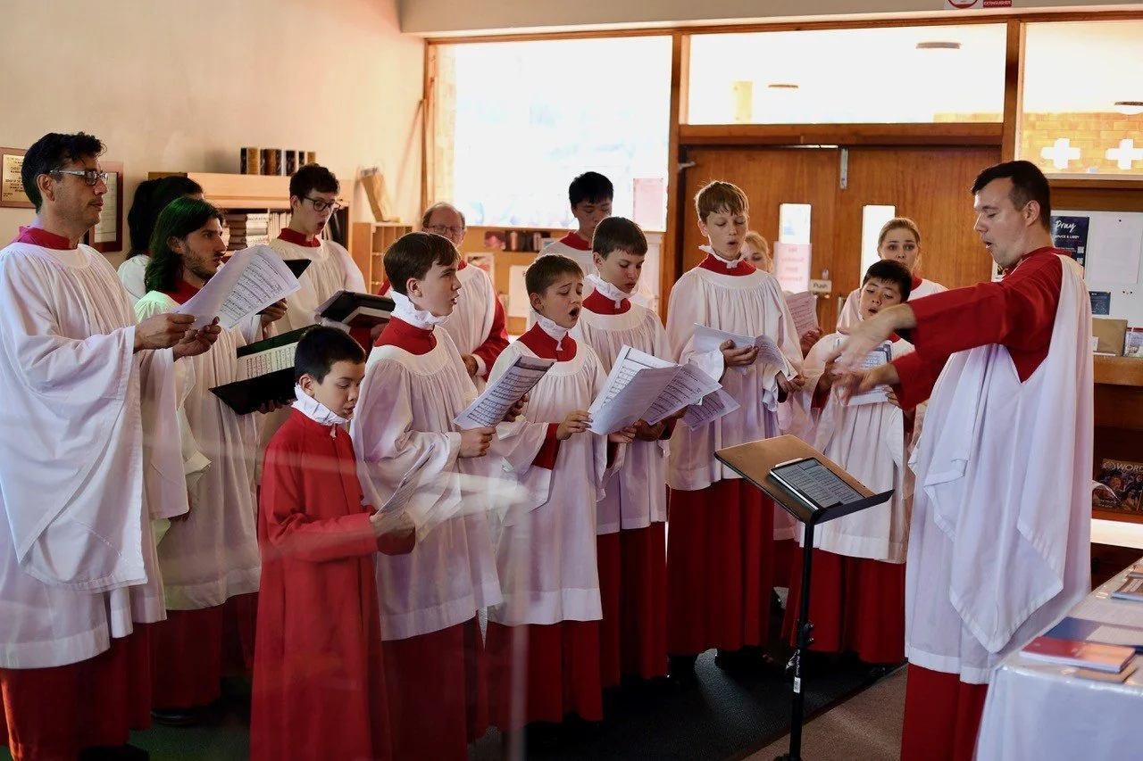 A church choir dressed in white robes with red accents singing during a service, led by a conductor in similar robes.