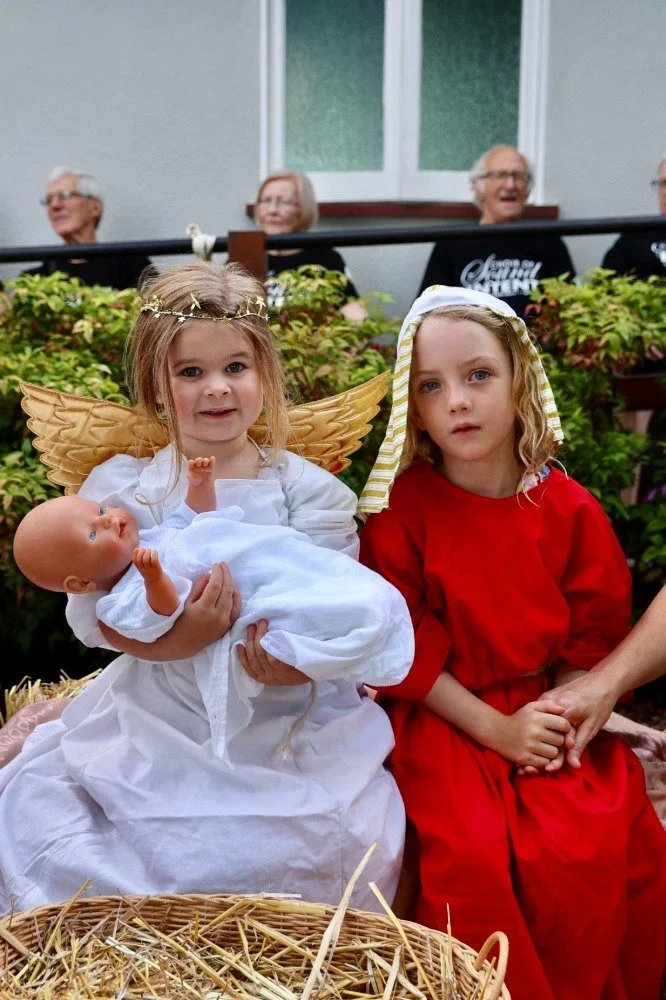 Children dressed as angel and Mary with a baby doll, participating in a nativity scene, with elderly people watching in the background.