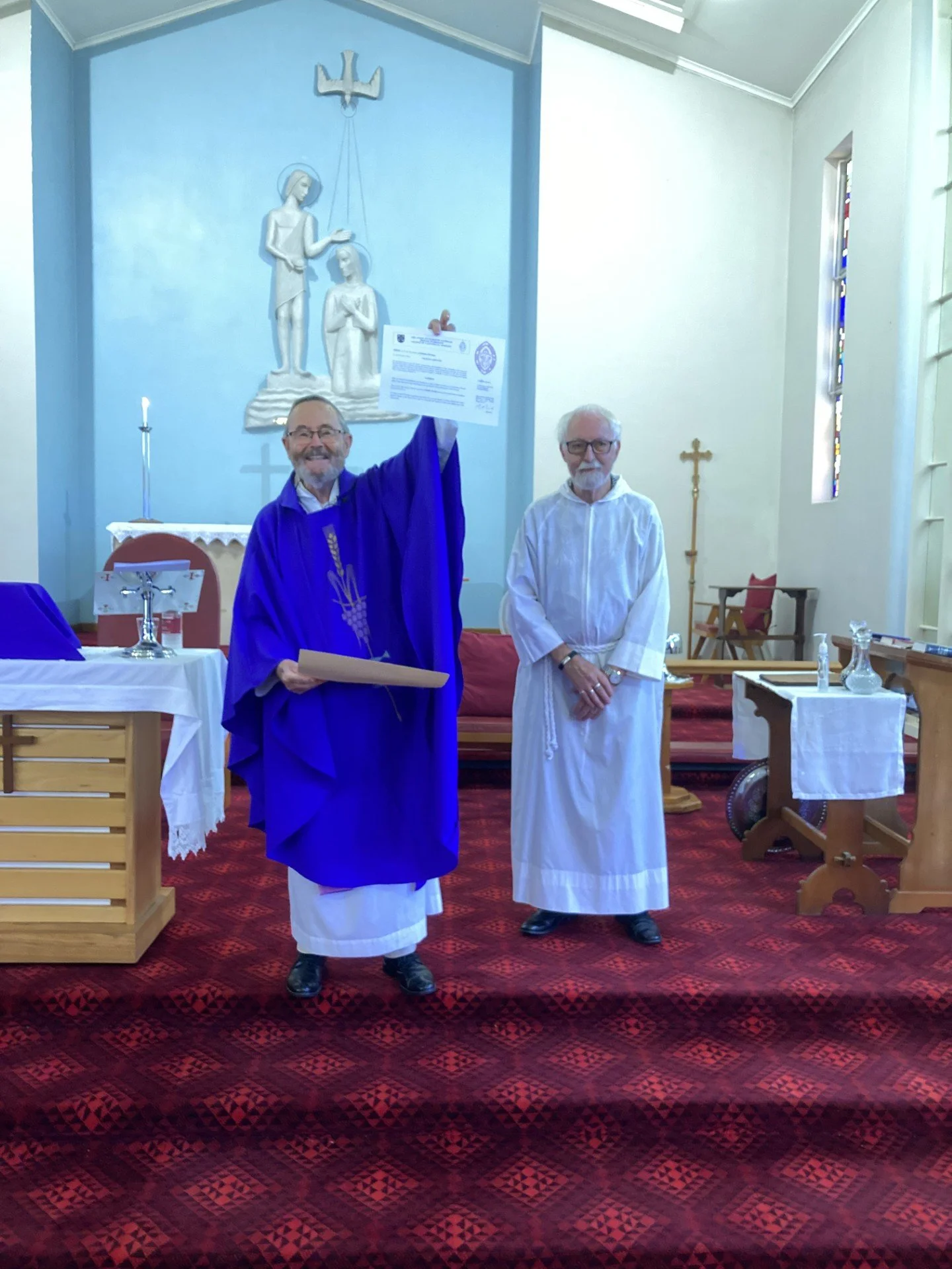 Two clergymen in church, one wearing a purple robe holding a certificate, the other in white robes standing beside him, with religious statues and symbols in the background.