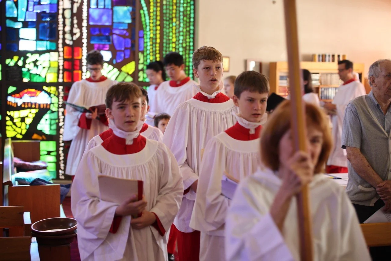 Young boys in white choir robes with red accents standing in line inside a church, likely during a religious service or procession, with colorful stained glass windows in the background.