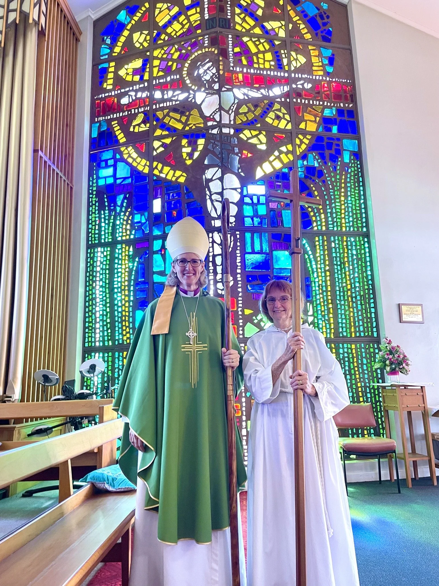 Two women dressed as Christian religious figures standing inside a church with a large, colorful stained glass window featuring a depiction of Jesus Christ on the cross in the background.