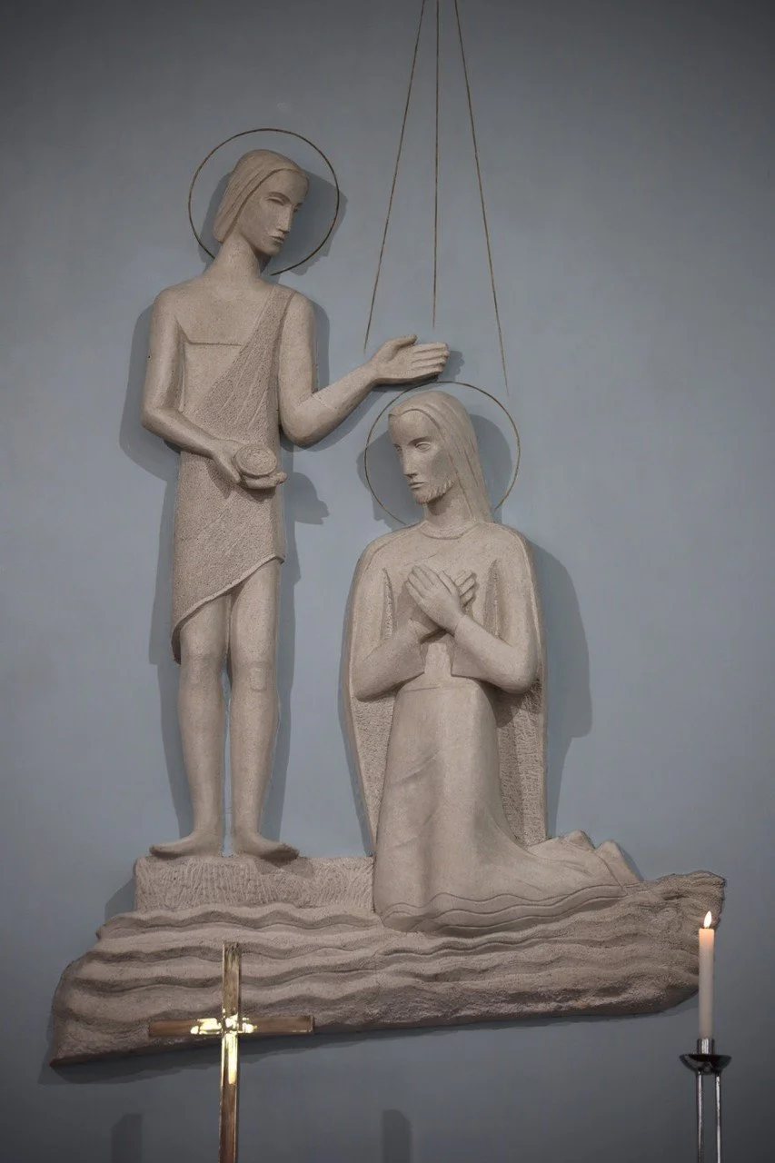 Stone sculpture of Jesus Christ kneeling with hands crossed on chest, receiving anointing from standing figure, in front of a plain background with a small candle and cross at the bottom.