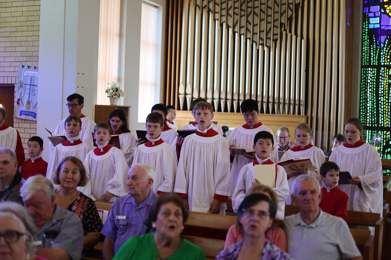 Children dressed in choir robes singing in a church with stained-glass windows and stained glass art on the walls, while congregation members sit in pews.