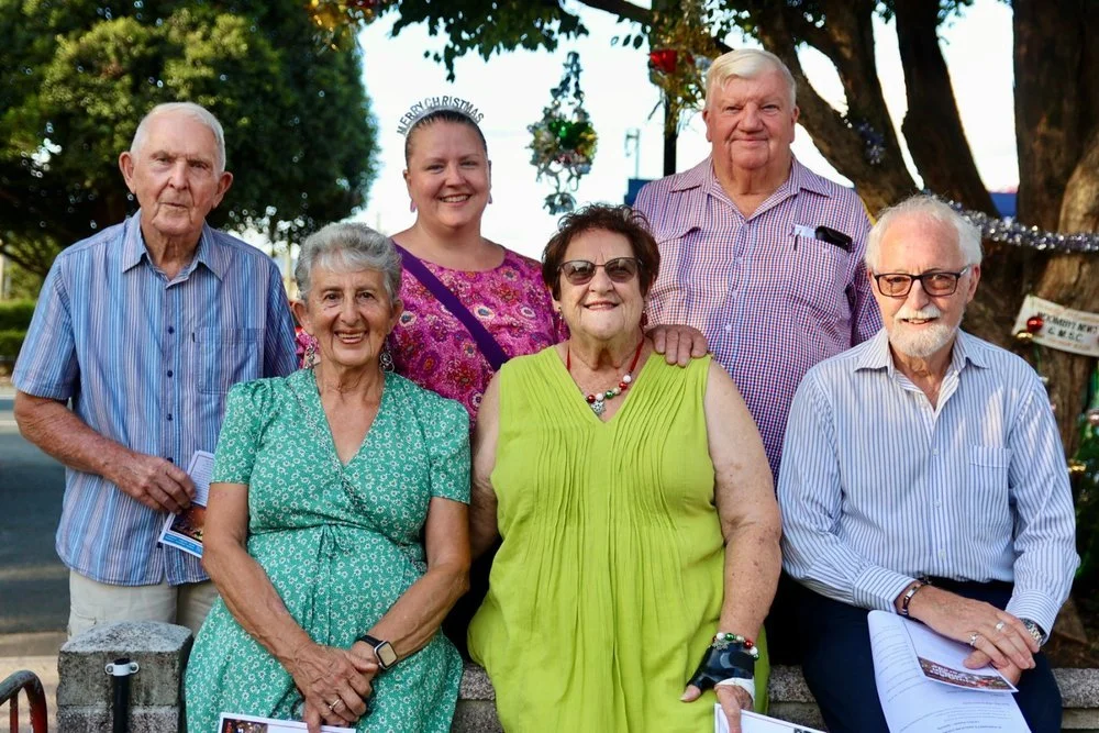 Group of seven senior adults posing outdoors, some wearing glasses and smiling, with Christmas decorations and trees in the background.