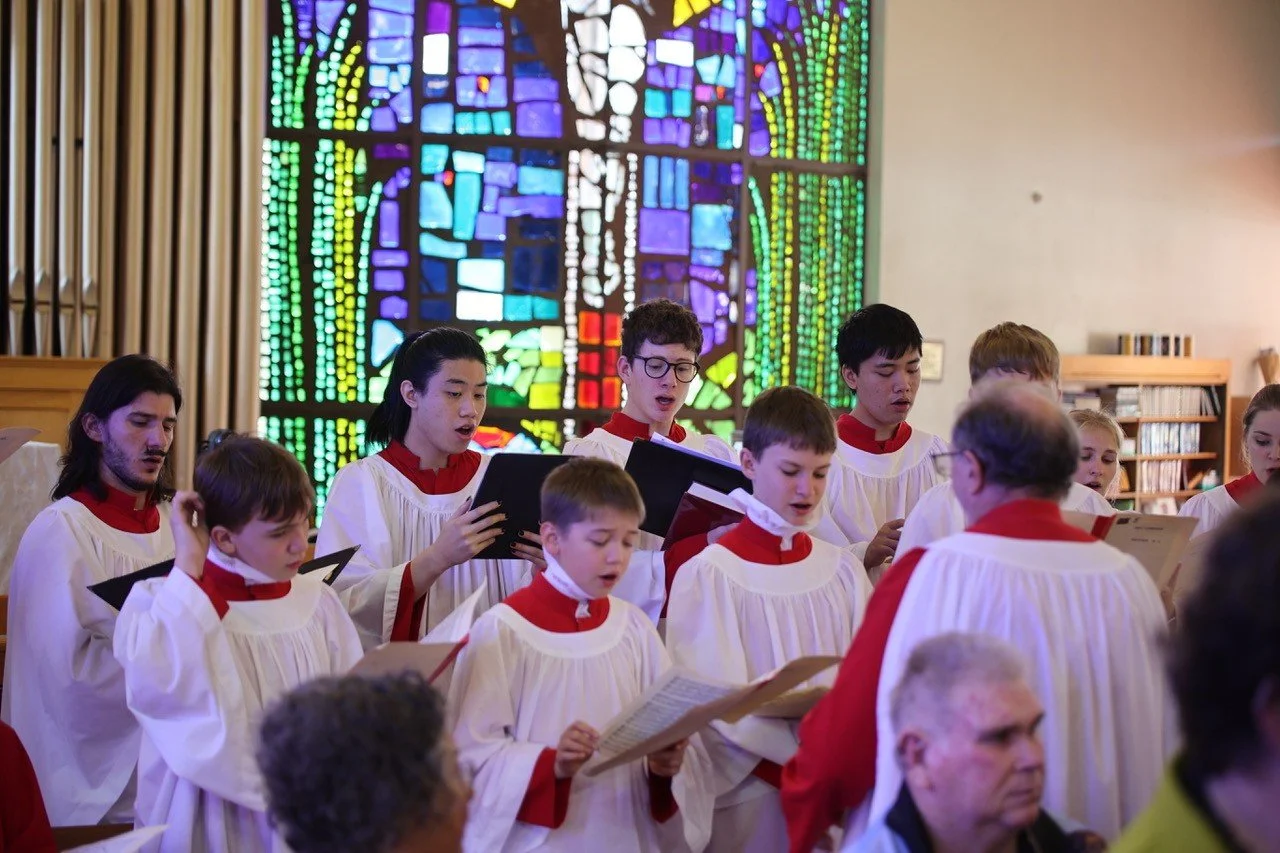A choir of children and teenagers singing in front of a colorful stained glass window inside a church.