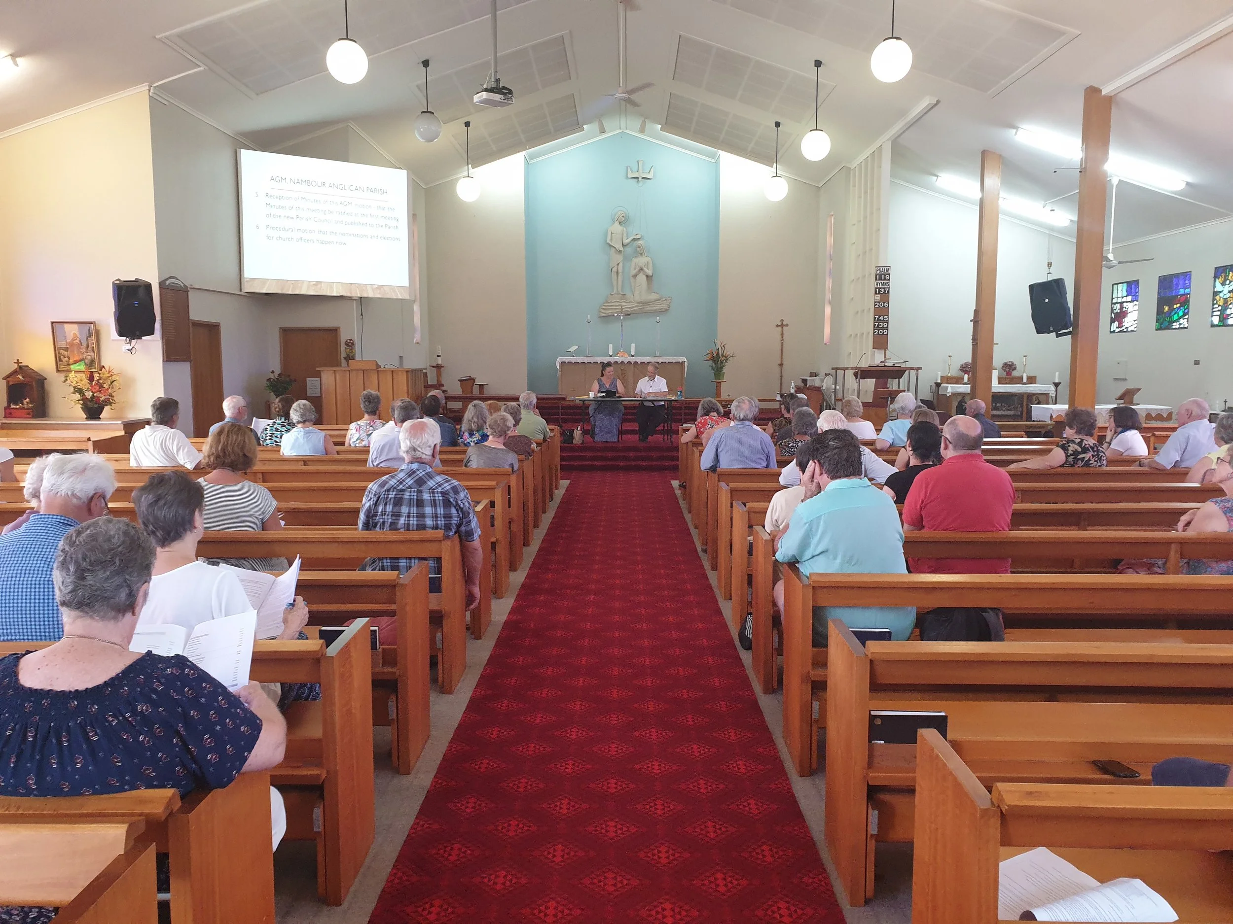 Inside a church with people sitting on wooden pews, facing the altar where two individuals are seated. The church has a high ceiling, hanging lights, a large crucifix, and stained glass windows.