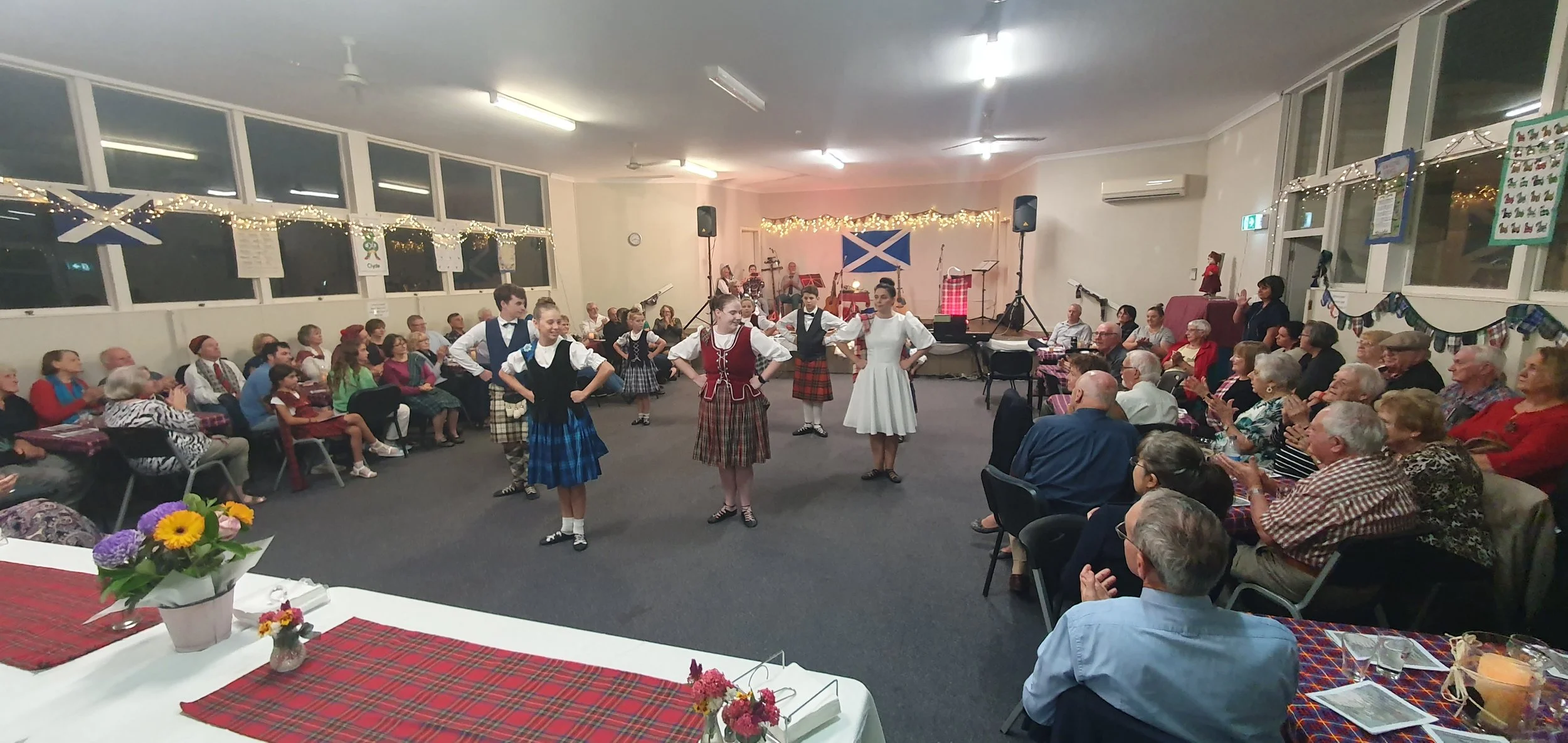 People gathered in a community hall watching children perform a traditional Scottish dance. The hall is decorated with Scottish flags, fairy lights, and festive banners, with an audience of all ages seated at tables.