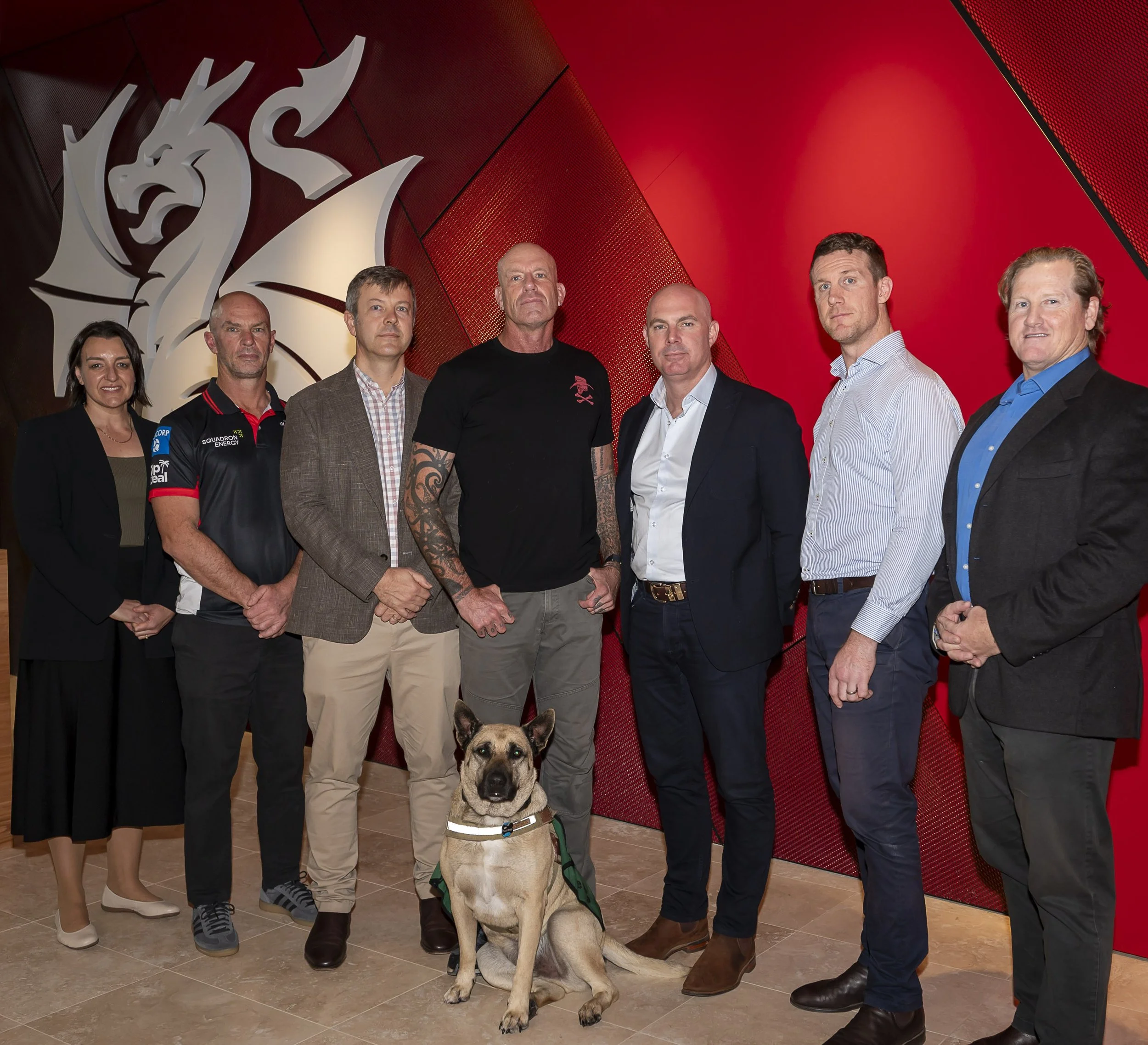 Group of seven people and a dog standing indoors against a red wall with a large white dragon sculpture. The group includes women and men dressed in business casual and sports attire, with various expressions.