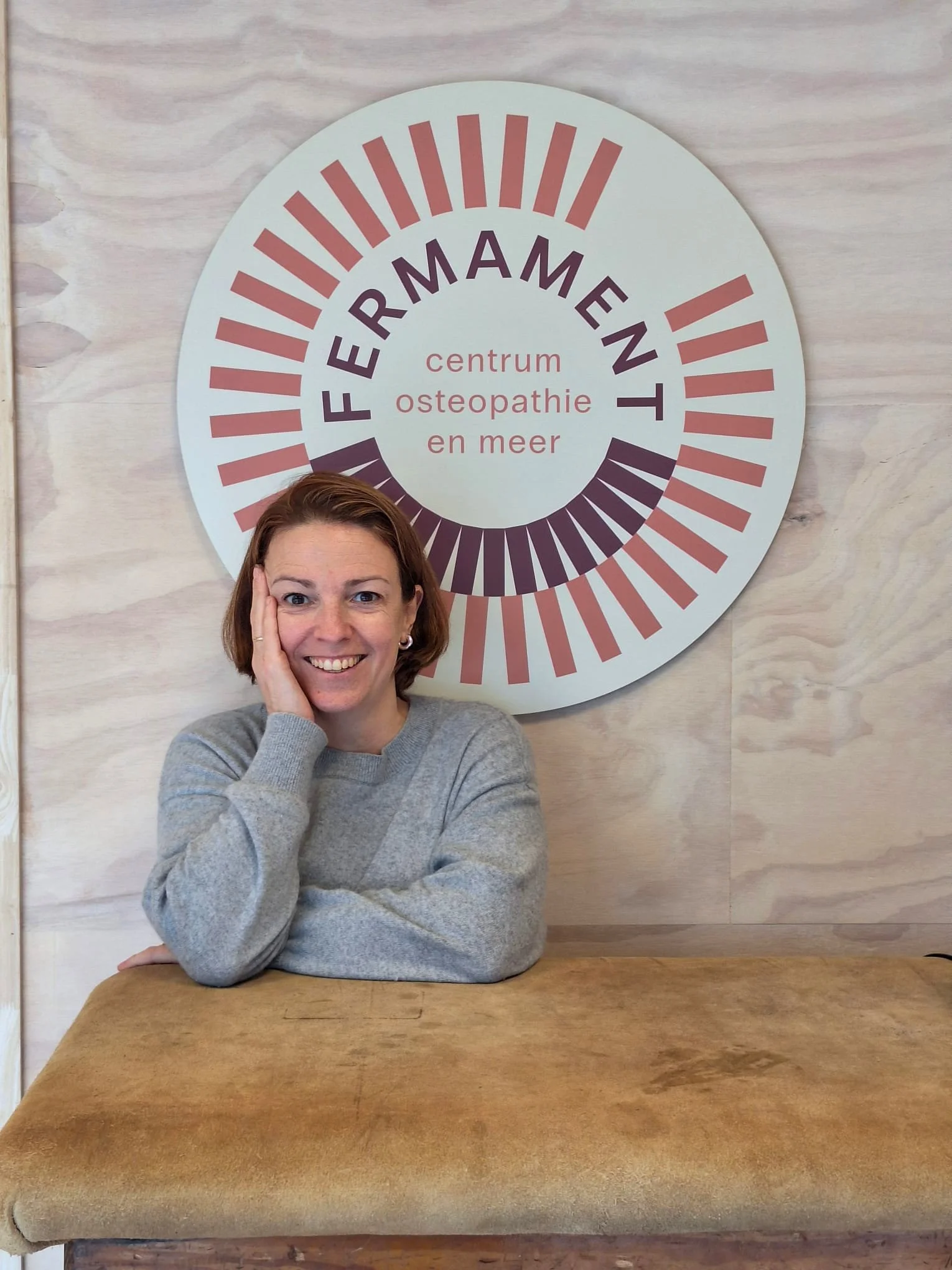 Een lachende vrouw met bruin haar en een grijs trui zit aan een houten tafel voor een muur met een groot cirkelvormig bord met de tekst 'FERRAMEN centrum osteopathie en meer'