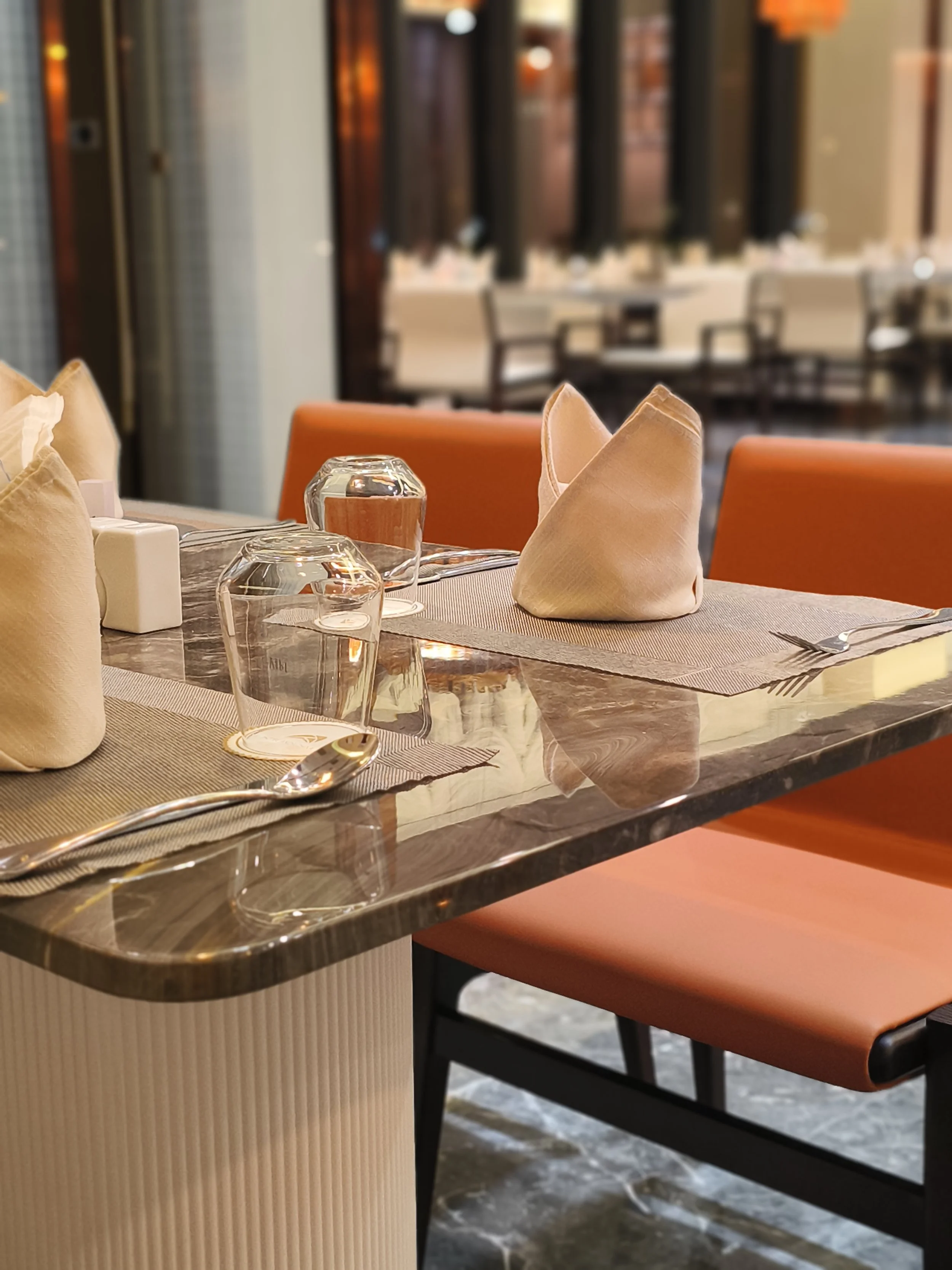A restaurant table with folded napkins, glasses, and cutlery, set for dining with orange chairs and a blurred background of additional seating.