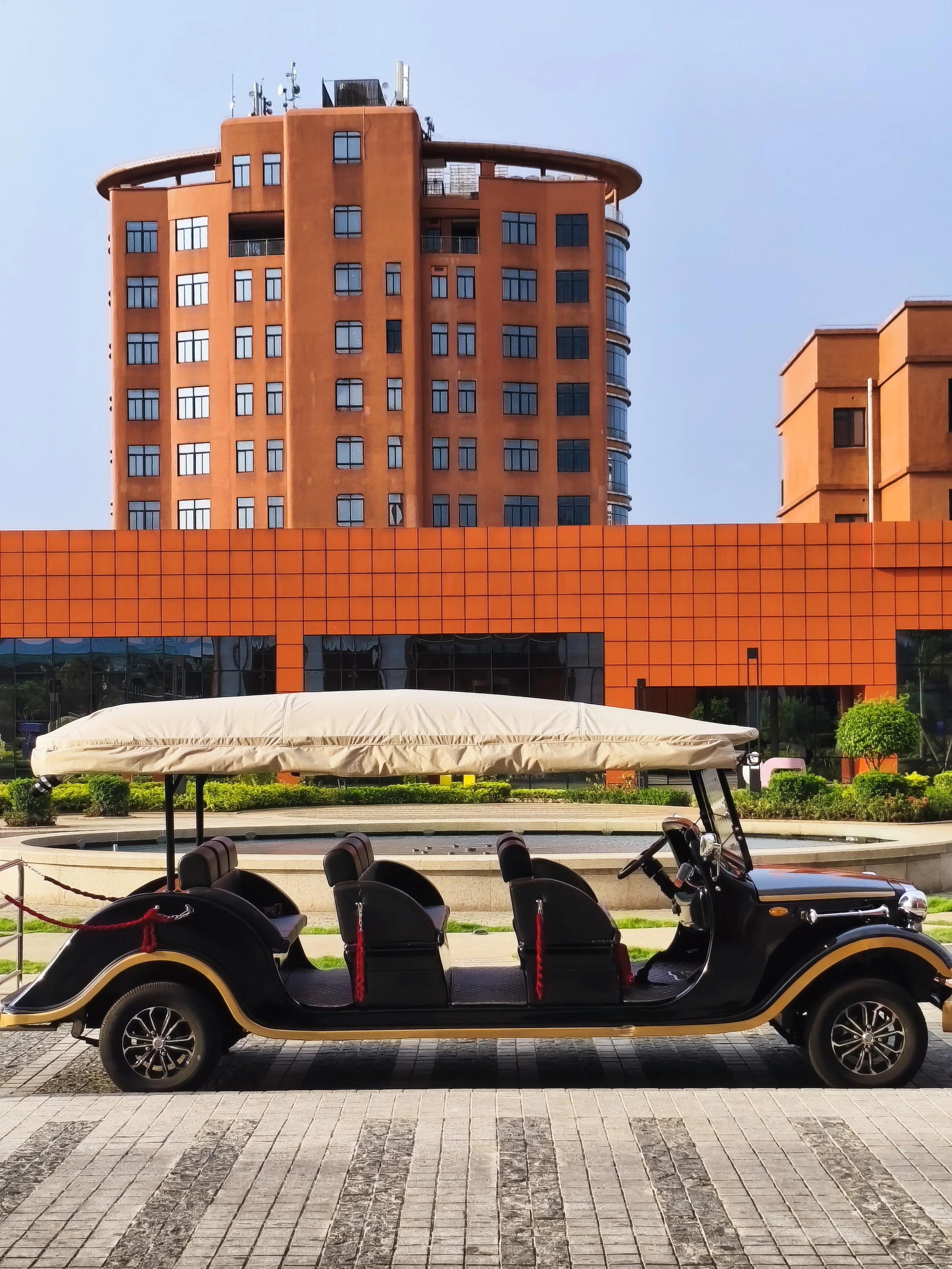 A black golf cart with four seats and a beige canopy parked on a paved area in front of a modern building with orange and brown exterior, large windows, and surrounding greenery.