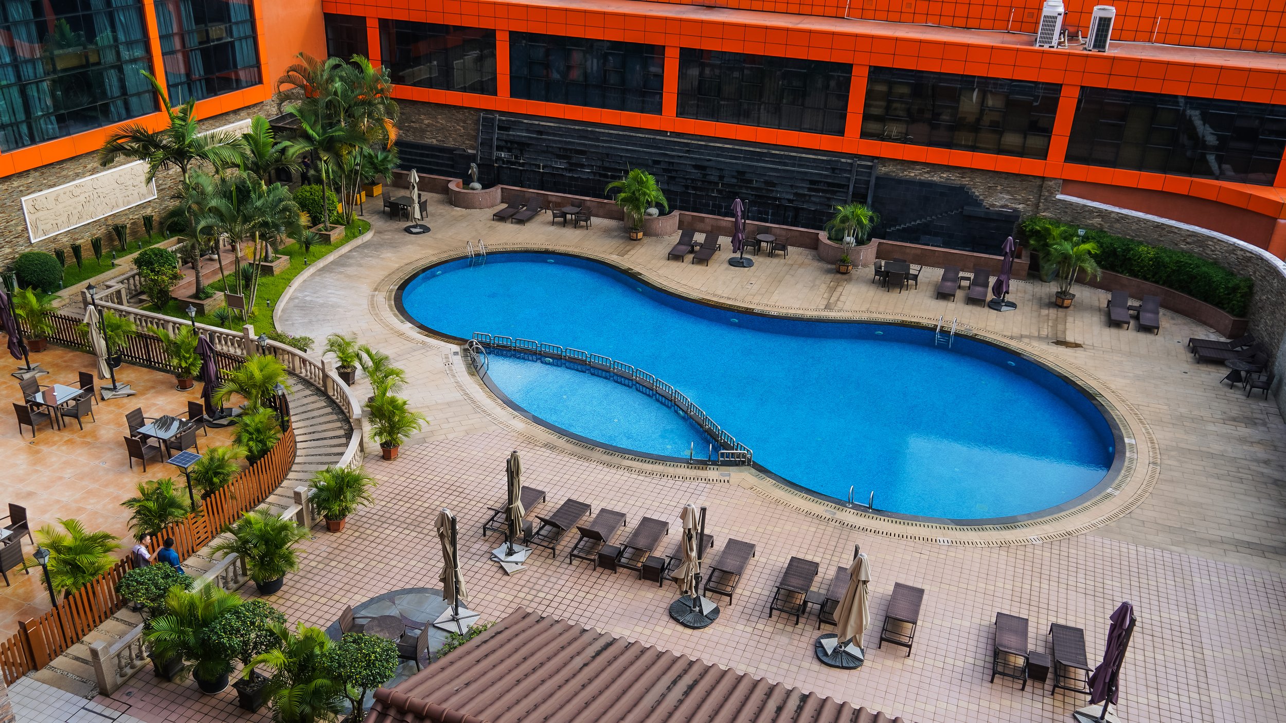 A hotel rooftop pool area with a kidney-shaped blue swimming pool, surrounded by lounge chairs, umbrellas, potted plants, and outdoor tables, with a modern orange building in the background.