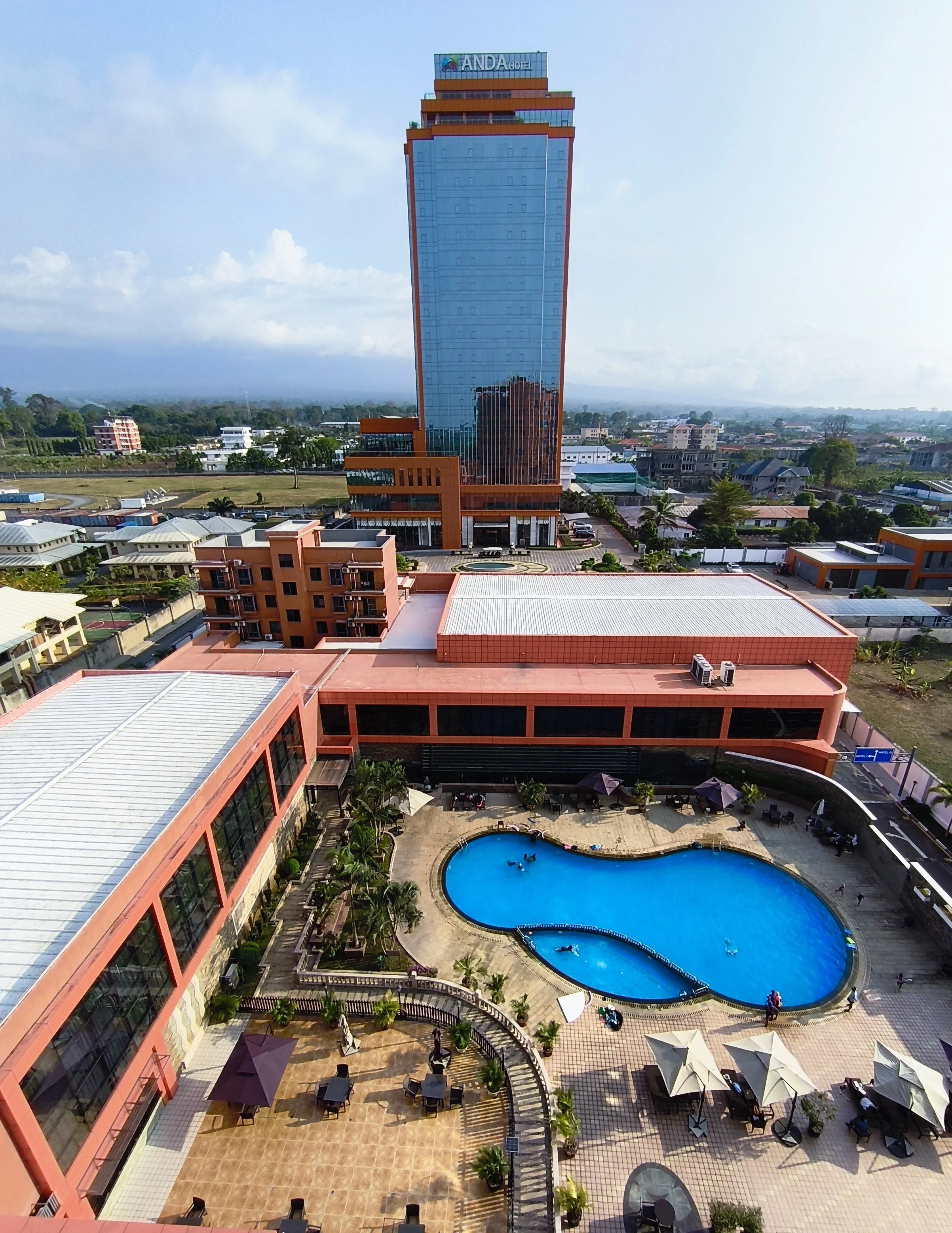 A tall hotel building with a glass facade, labeled 'ANDA HOTEL,' overlooking a swimming pool with a deck and umbrellas, surrounded by smaller buildings and greenery under a partly cloudy sky.