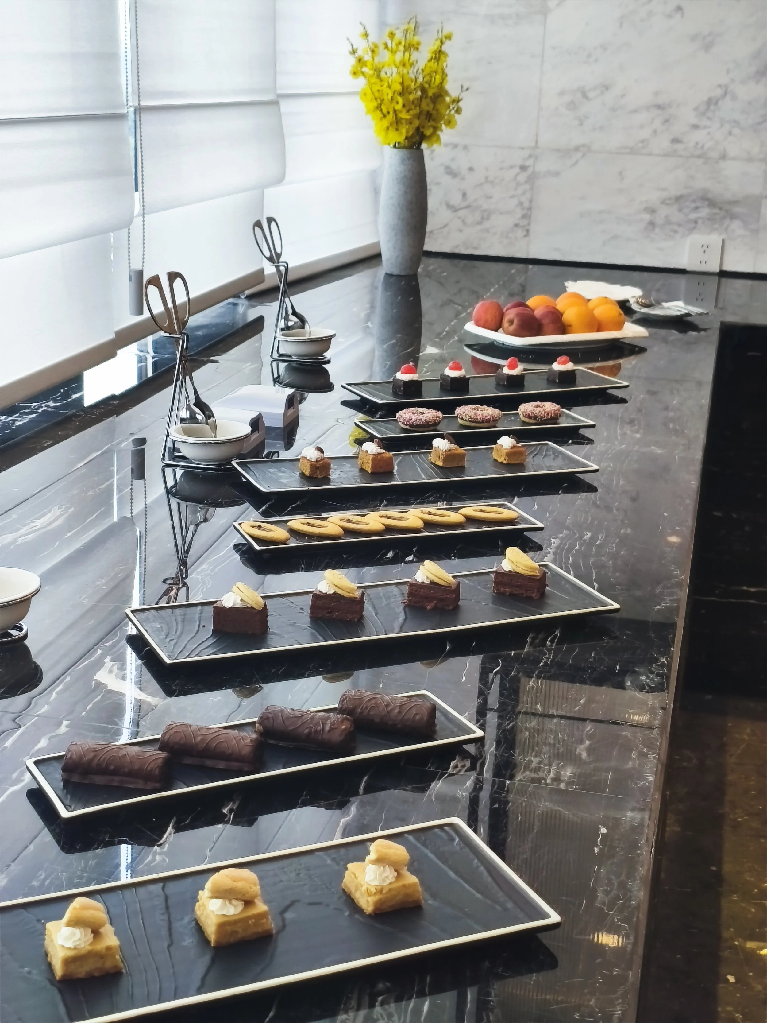 An array of desserts on black rectangular plates arranged on a marble counter in a modern kitchen with white window blinds, a gray vase with yellow flowers, and a fruit platter in the background.