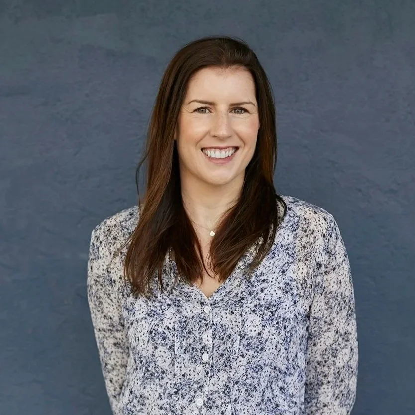 Meet Anthea Barry, A woman with brown hair smiling, wearing a long-sleeve floral blouse, standing against a blue wall.