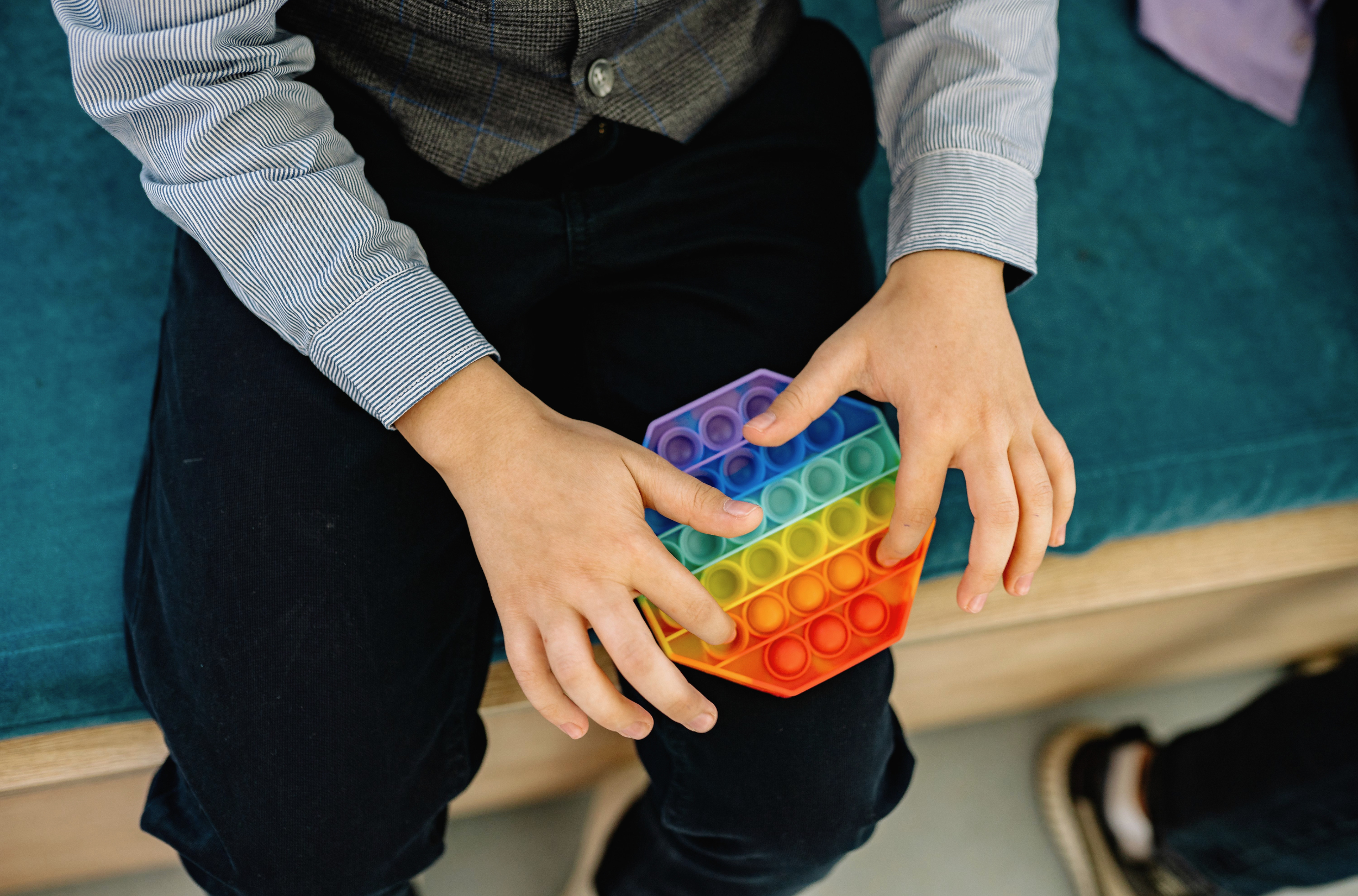 A child in a striped shirt and dark vest sits on a teal bench, pressing the bubbles of a rainbow-coloured pop-it fidget toy held in their lap.
