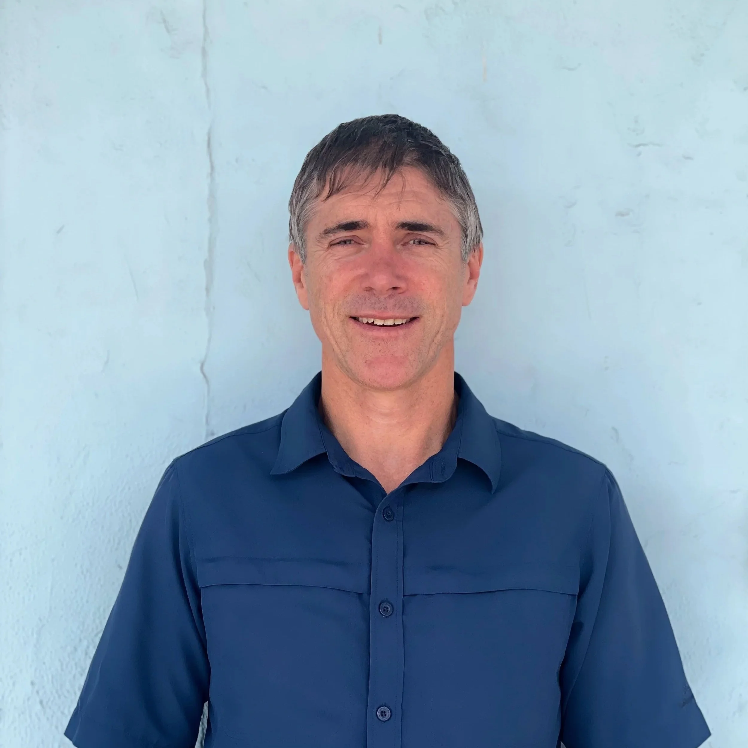 A man with short gray hair and a slight smile, wearing a navy blue collared shirt, standing against a light blue wall.