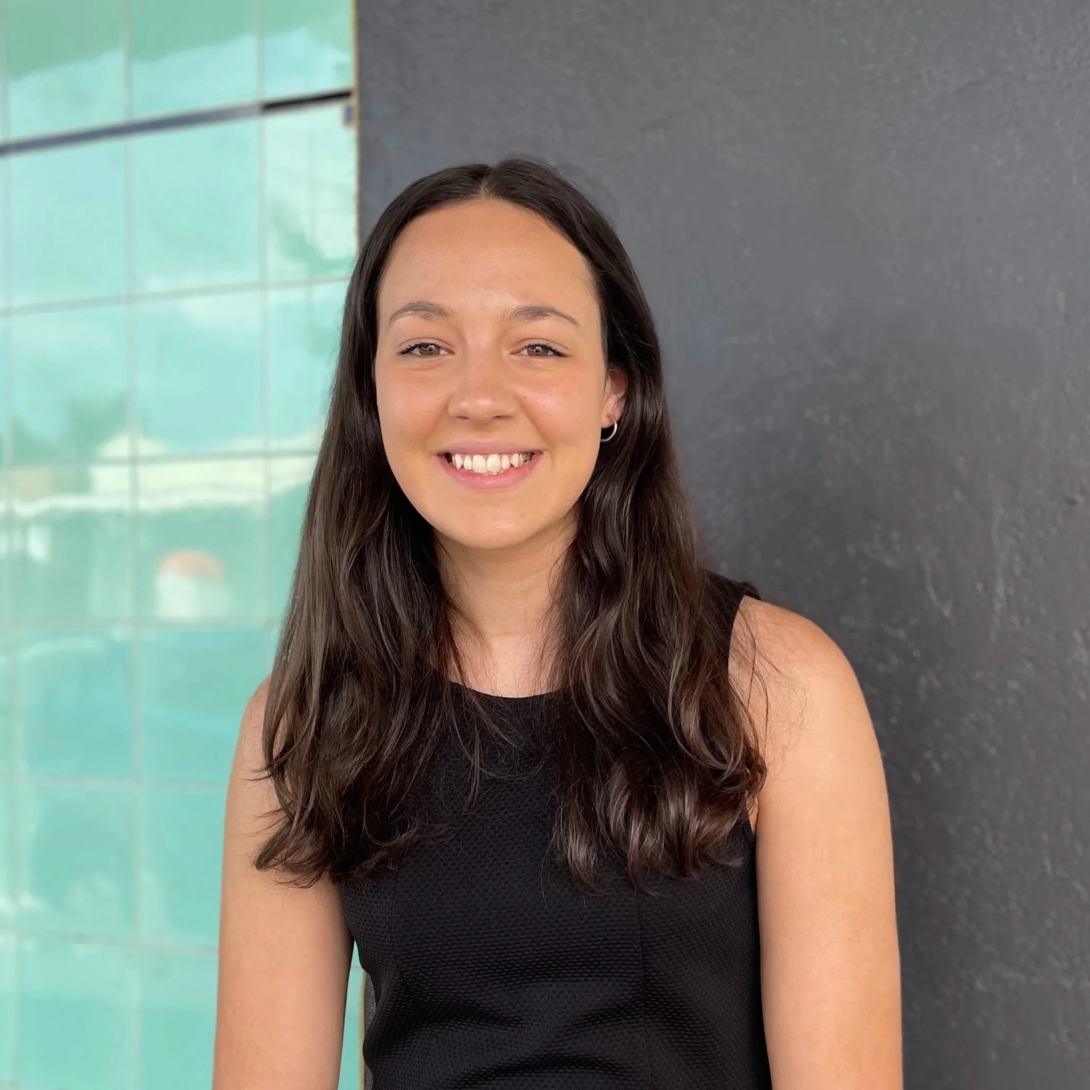 Meet Tiahn Biundo, A young woman with long dark hair wearing a sleeveless black top, smiling, standing outdoors in front of a dark wall and a glass window.
