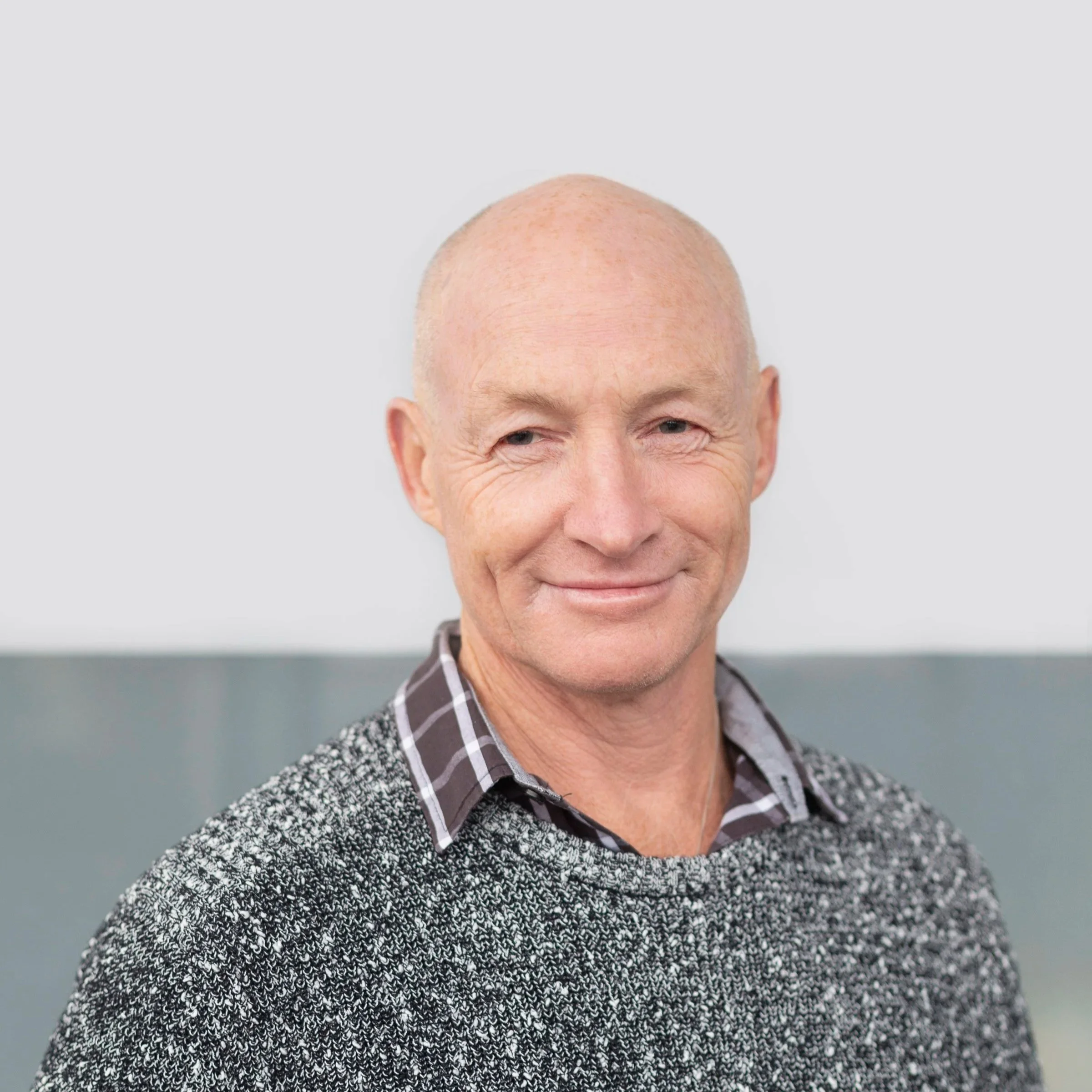 Meet Ian Connop, A smiling bald man in a checkered shirt and gray sweater standing against a plain background