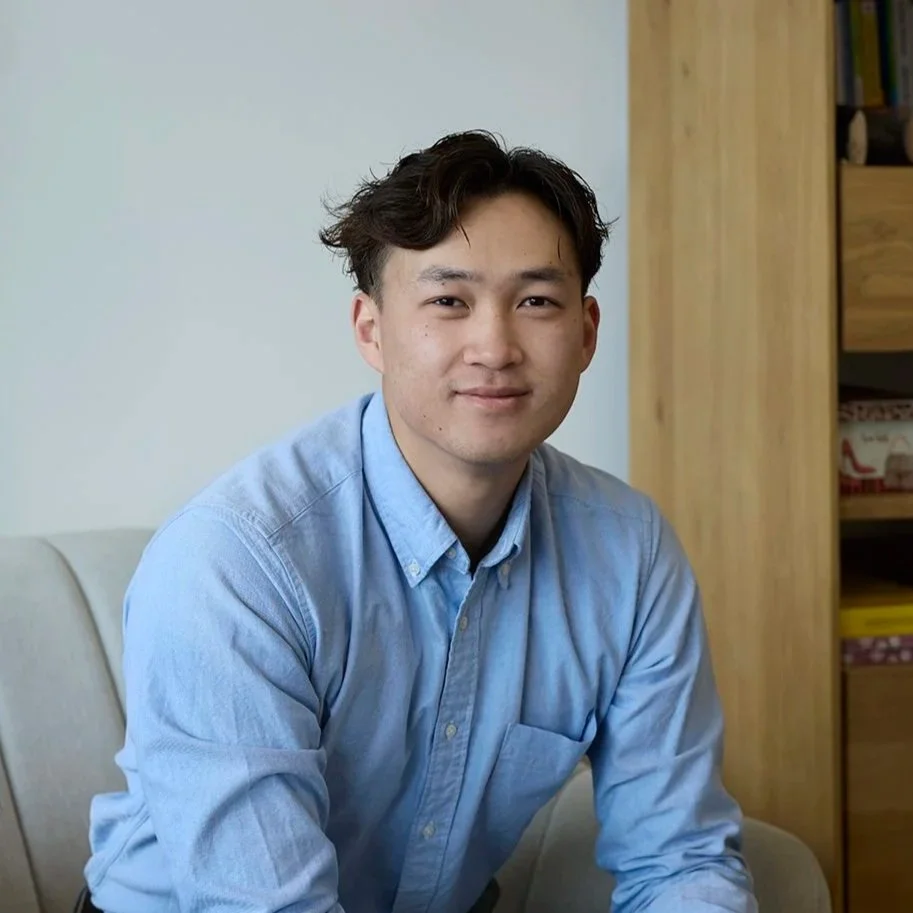 A young man with dark hair in a light blue button-up shirt sitting on a beige couch in a living room, smiling at the camera. There is a pink vase with dried flowers on the armrest and wooden bookshelves in the background.