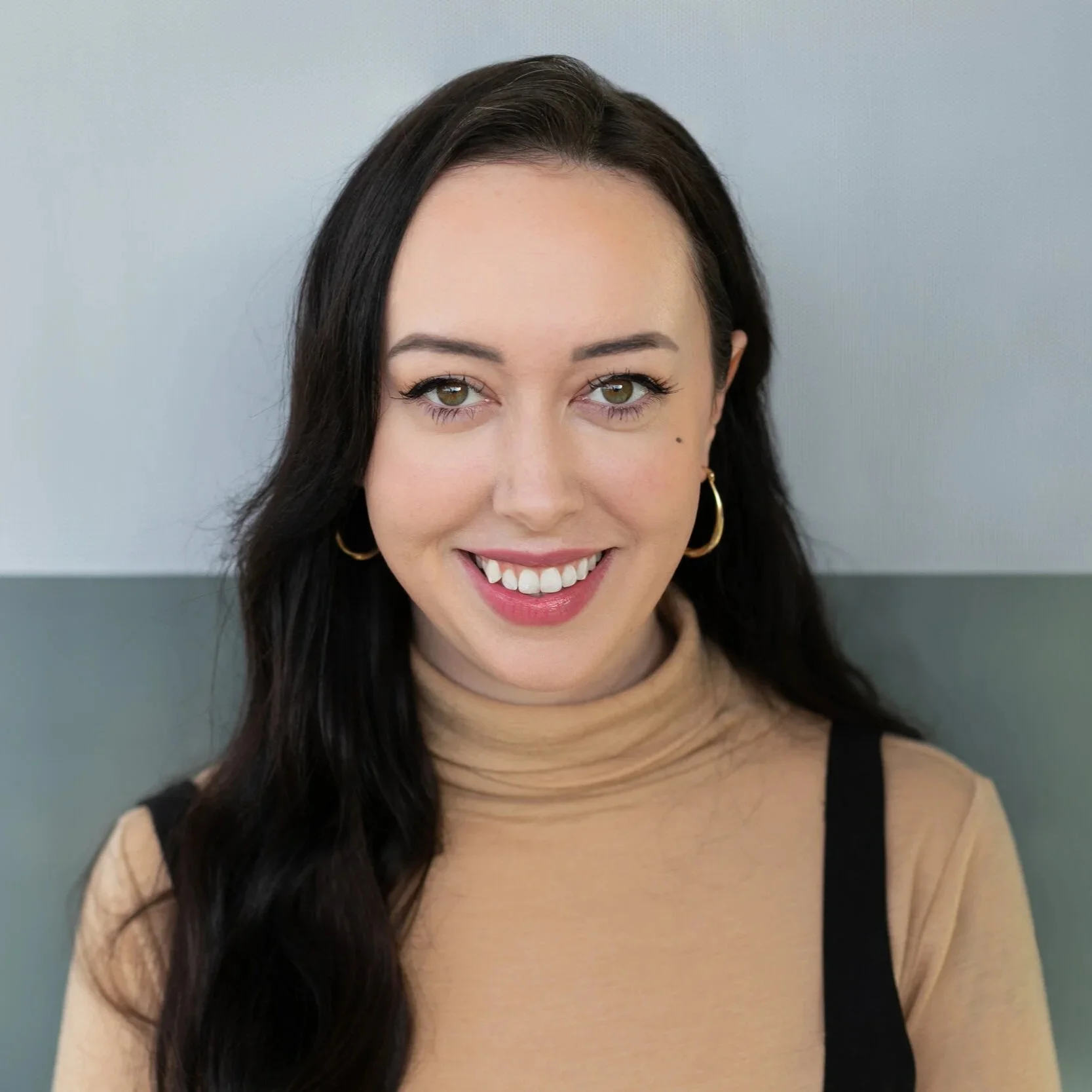 Meet Roni Sokolic, A smiling woman with long dark hair, wearing a beige turtleneck and black straps, standing against a two-tone background.