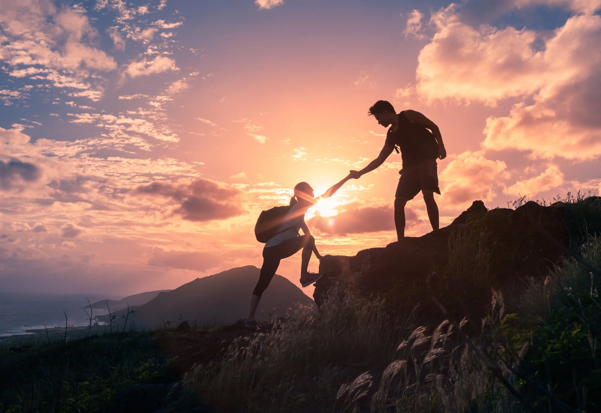 Two hikers, a man and a woman, helping each other climb a rocky hill at sunset with a scenic landscape in the background.