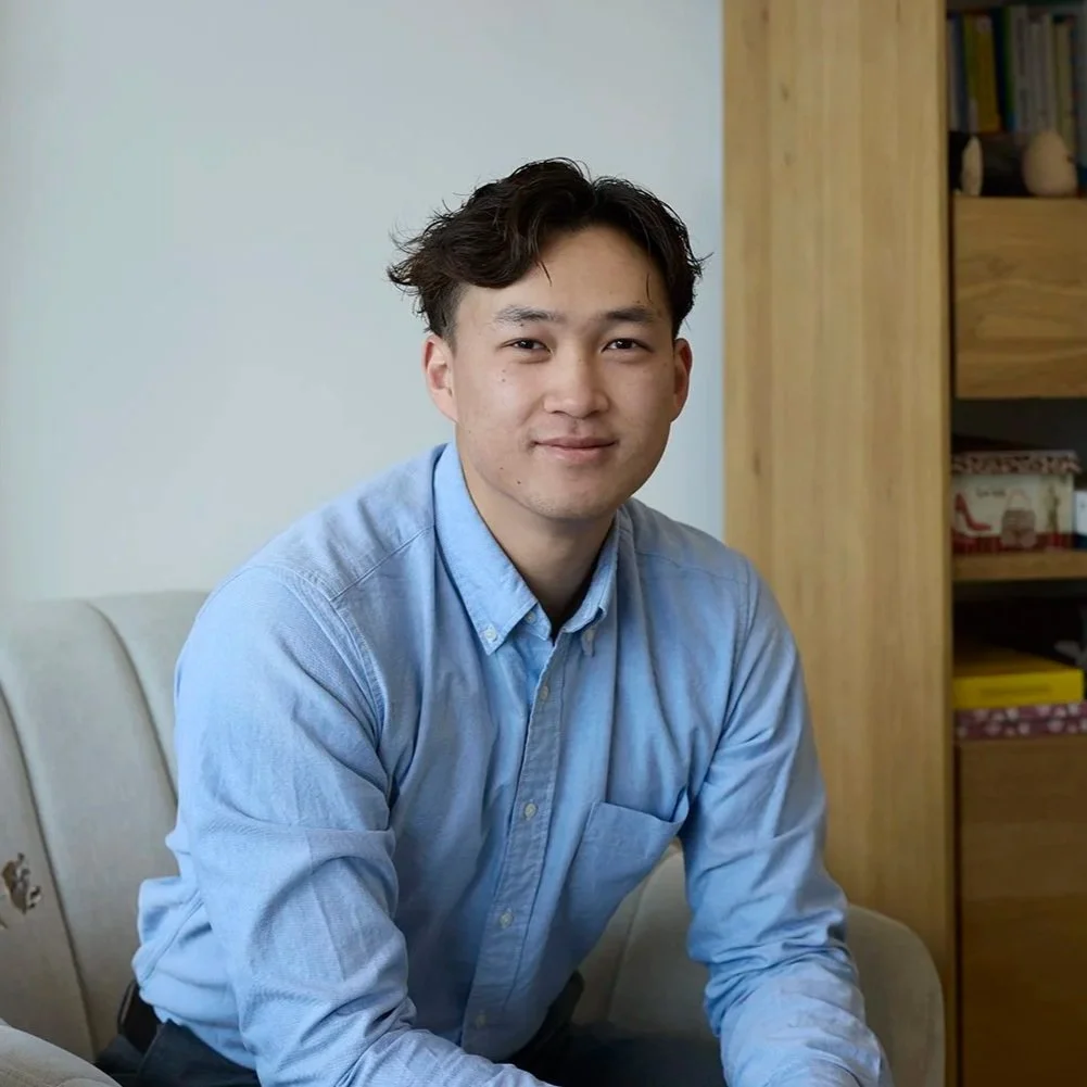 A young man with dark hair in a light blue button-up shirt sitting on a beige couch in a living room, smiling at the camera. There is a pink vase with dried flowers on the armrest and wooden bookshelves in the background.