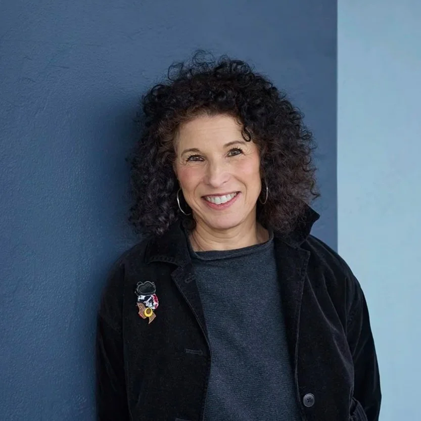 A woman with curly dark hair smiling, standing against a blue wall, wearing a black jacket with a pin, hoop earrings, and a grey shirt.
