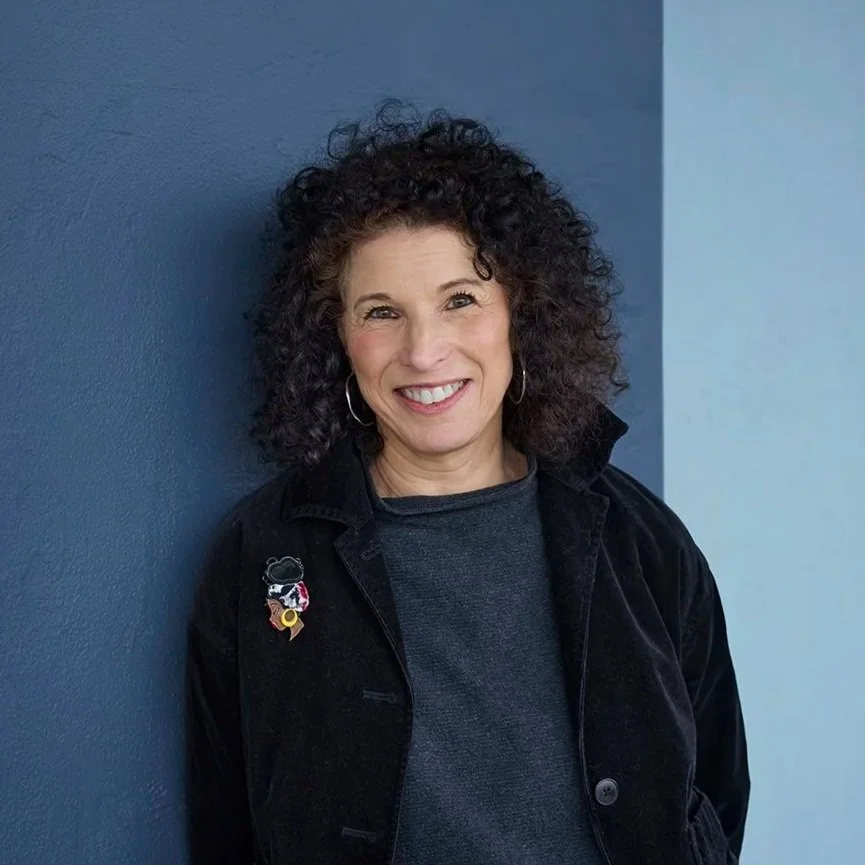 A woman with curly dark hair smiling, standing against a blue wall, wearing a black jacket with a pin, hoop earrings, and a grey shirt.