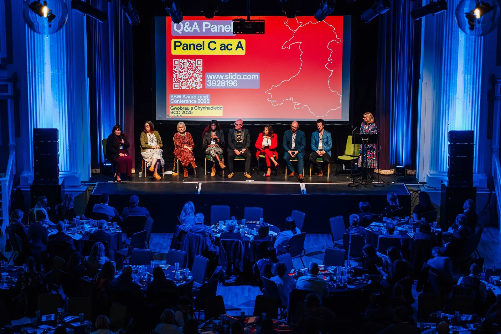 A panel of nine people seated on stage with a woman at a podium to the right, in front of a large red screen displaying text and graphics, surrounded by an audience in a conference setting.