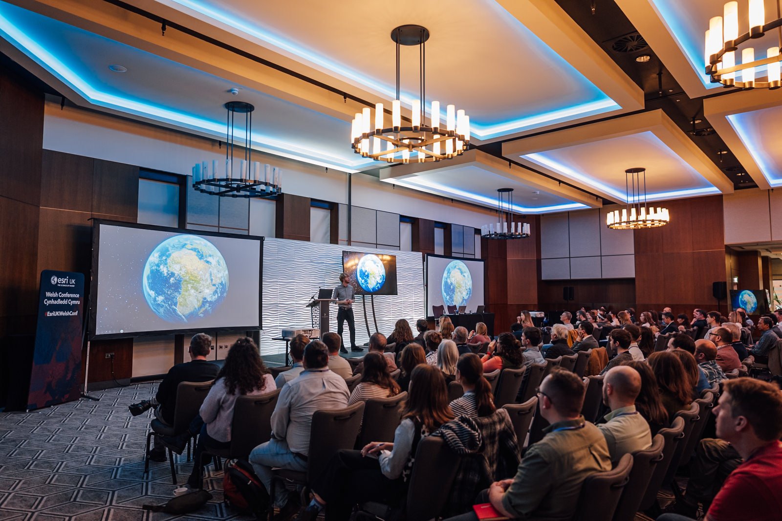A conference room filled with seated attendees watching a speaker on stage. The speaker stands in front of large screens displaying images of Earth. The room has modern lighting and decor.