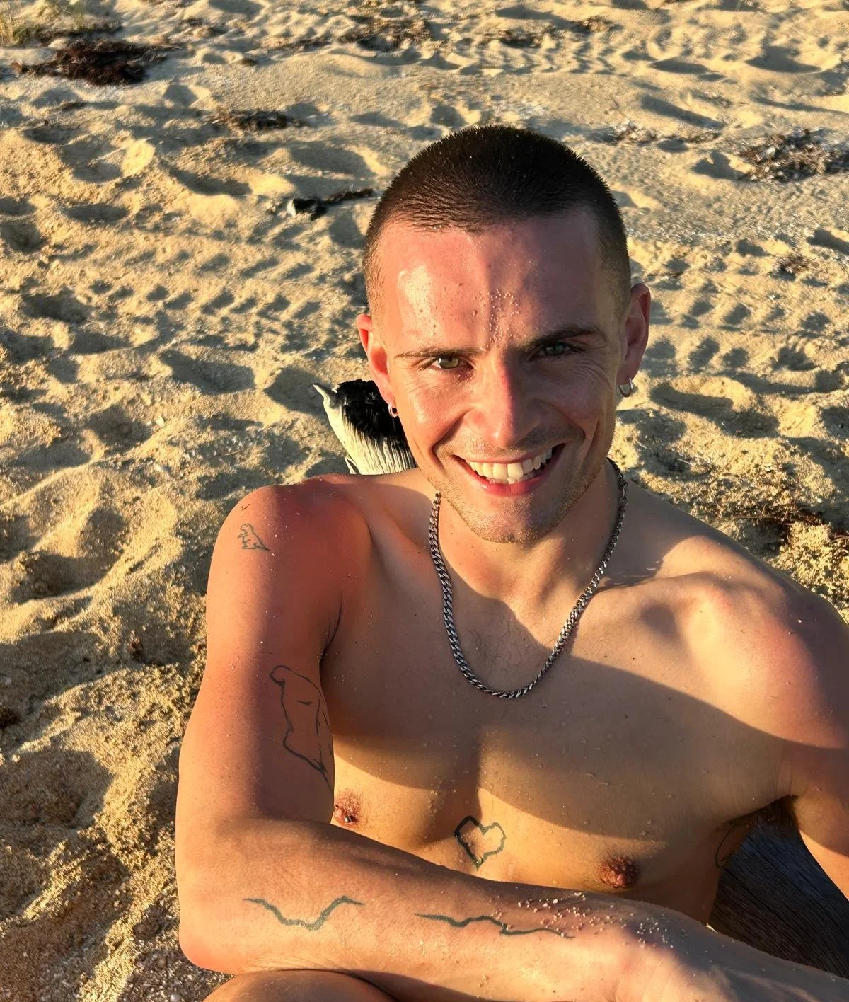 A smiling young man with tattoos taking a selfie on the beach, with sand and seaweed in the background.