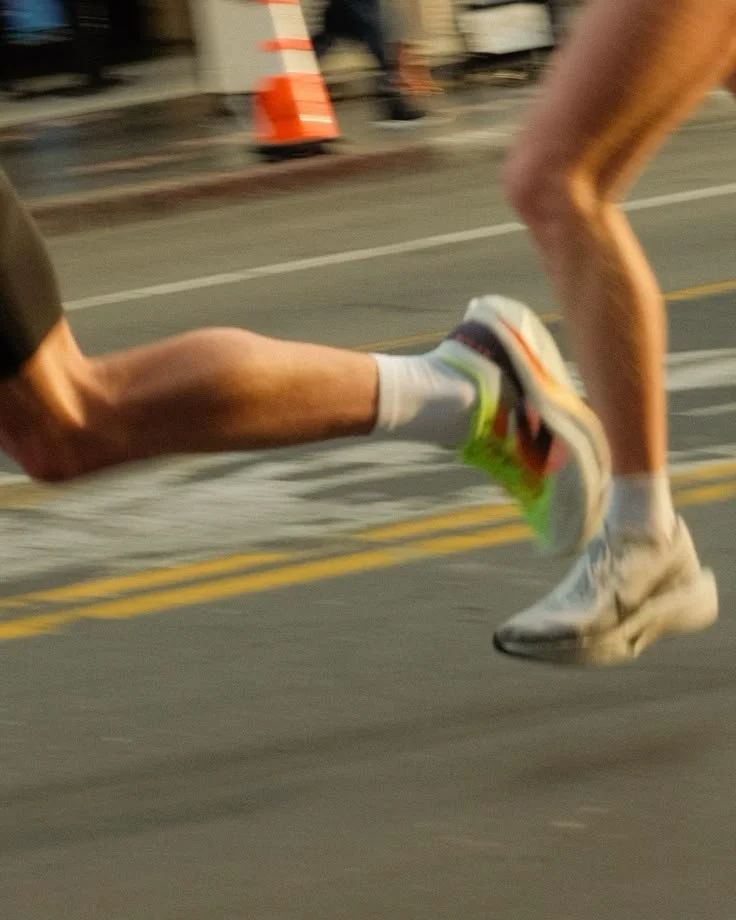 Close-up of two runners' legs as they sprint on a street, with one runner's foot just off the ground.