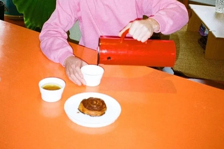 Person wearing a pink shirt pouring hot water from a red kettle into a white cup on an orange table, with a plate holding a cinnamon roll and a cup of coffee nearby.