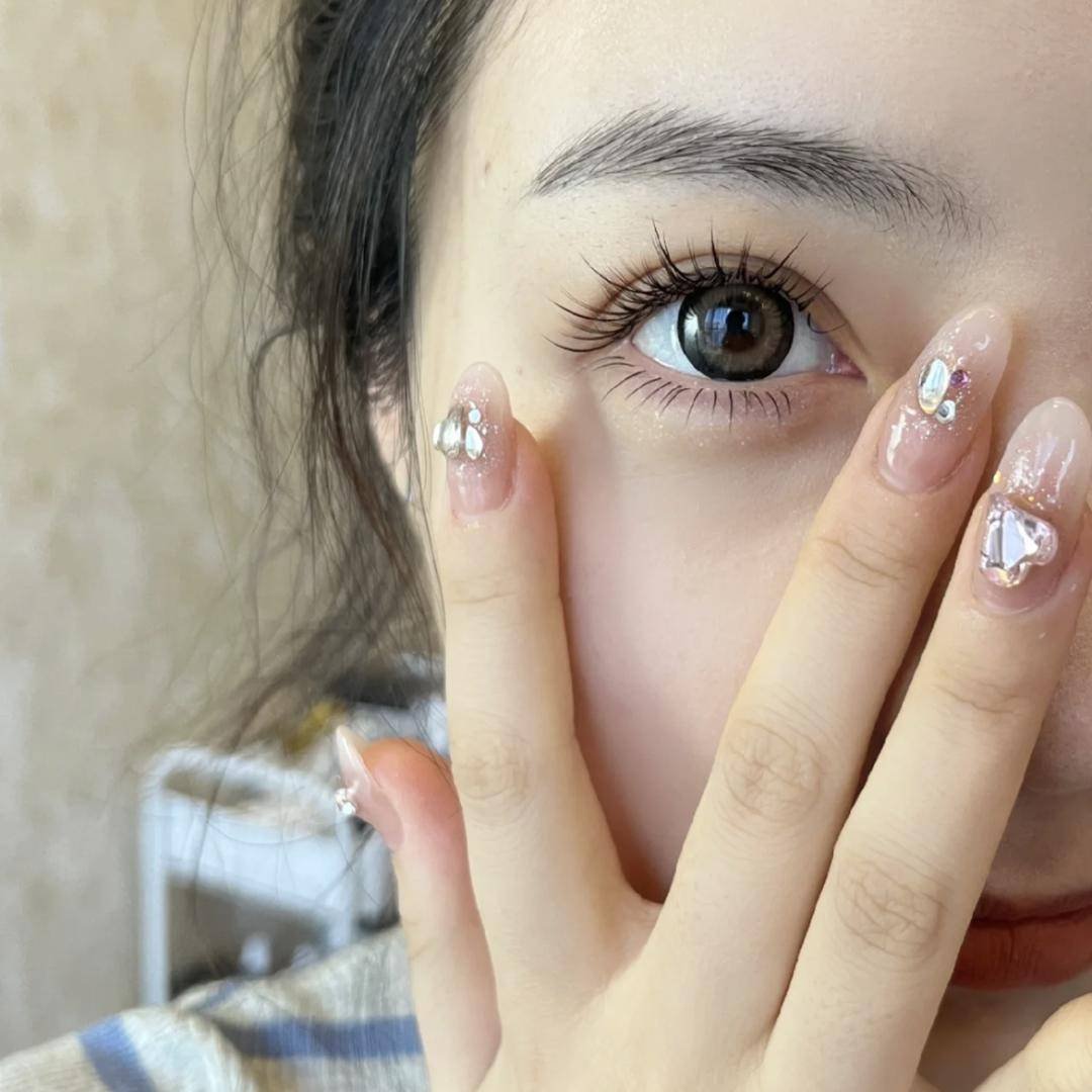 Close-up of a woman's eye with elaborate nail art featuring rhinestones and intricate decorations on her fingers