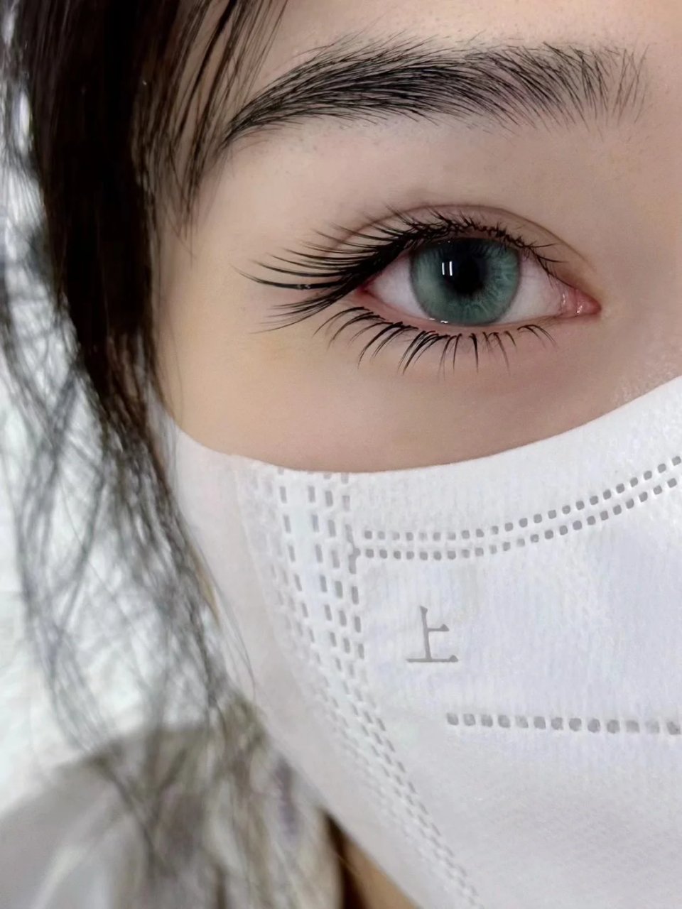 Close-up of a person's eye with long lashes, wearing a white face mask, showing part of their eyebrow.