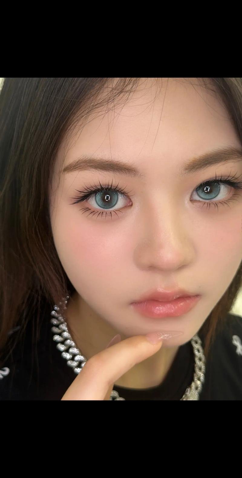 Close-up portrait of a person with blue eyes, long eyelashes, and light makeup, wearing a silver necklace.