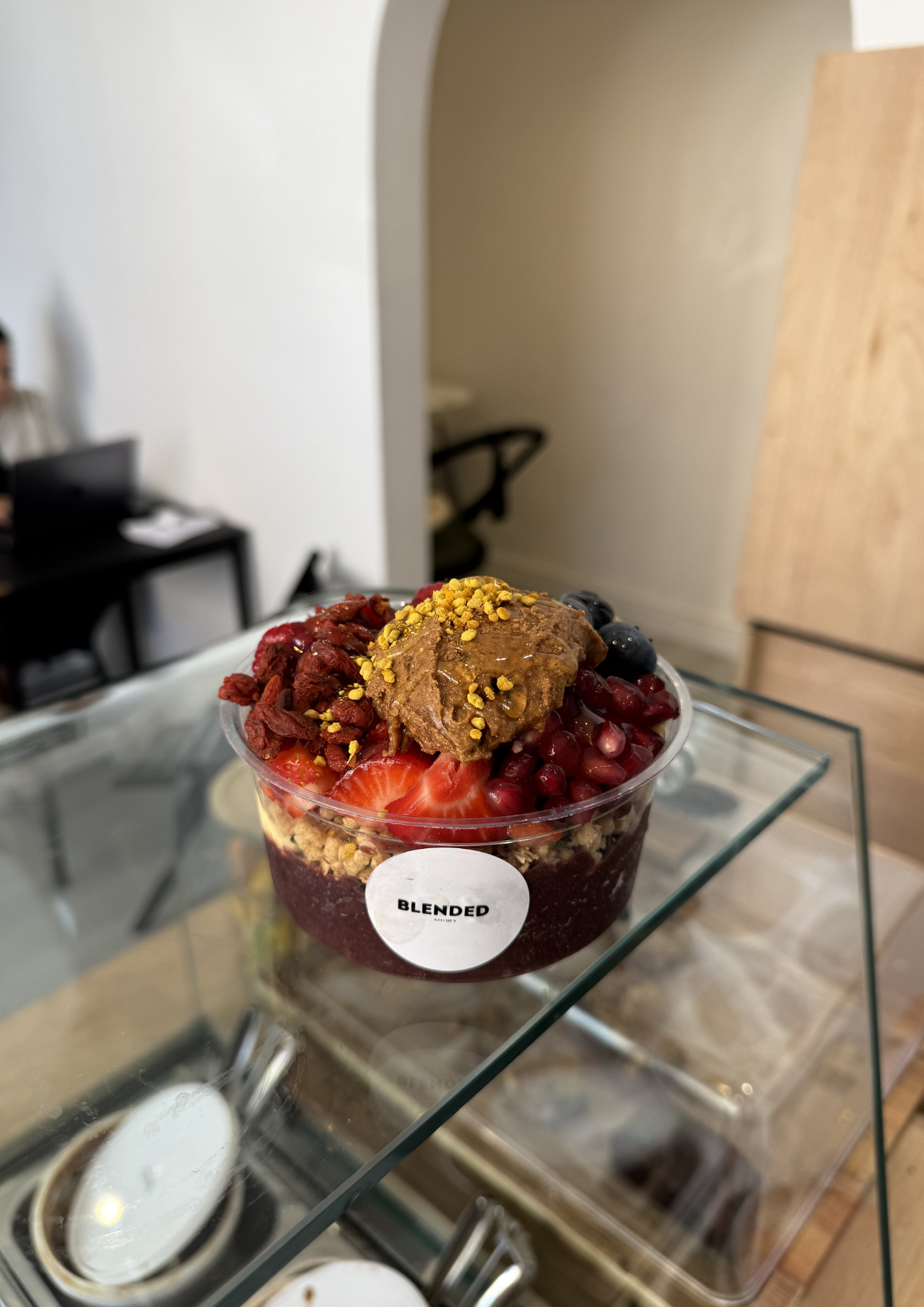 A cup of mixed berries topped with granola and a cookie on a glass table, with a blurred background of a dining area.