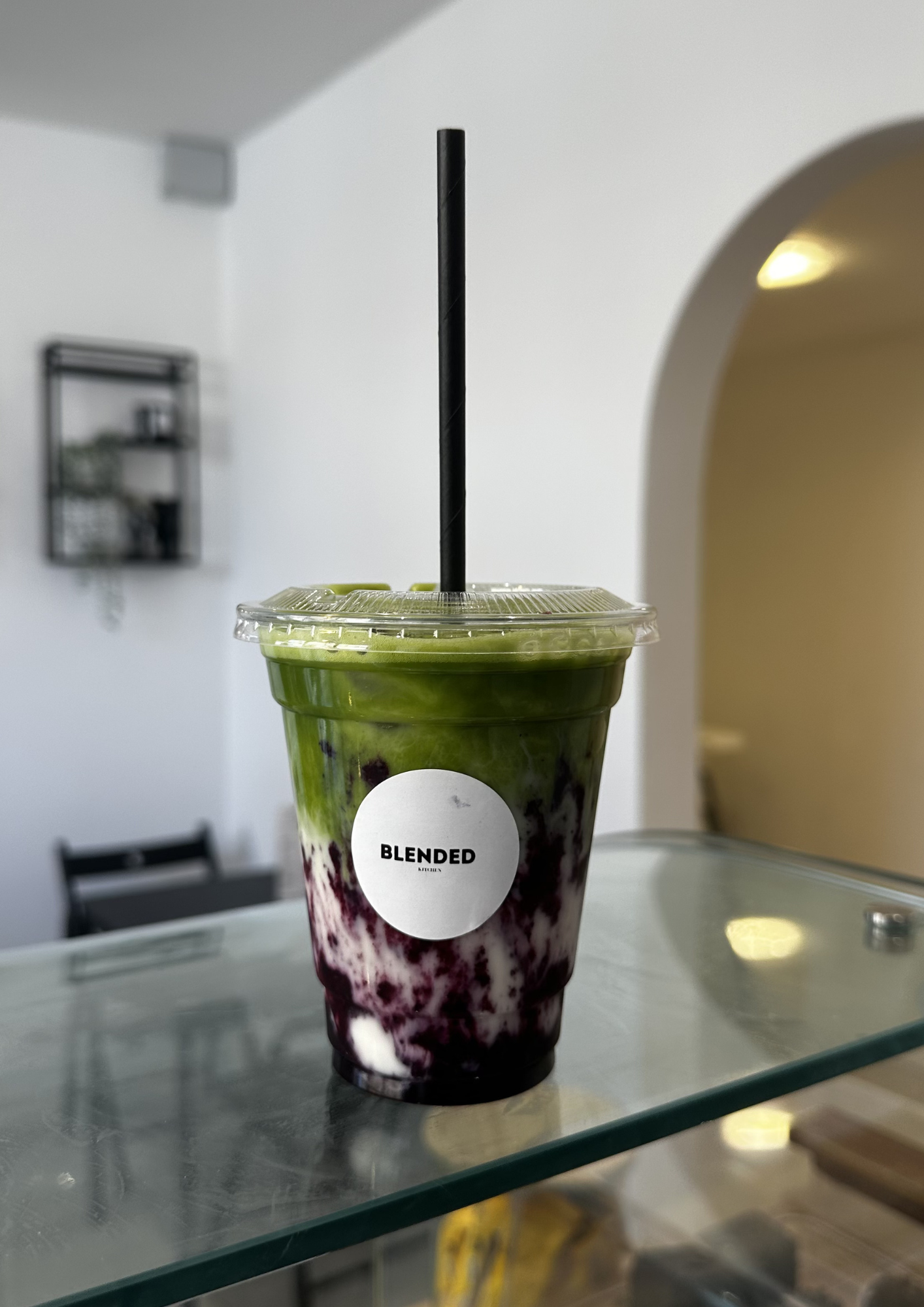 A plastic cup of blended smoothie with blueberries, green pineapple, and purple dragon fruit, labeled 'BLENDED,' on a glass counter inside a cafe or juice bar.