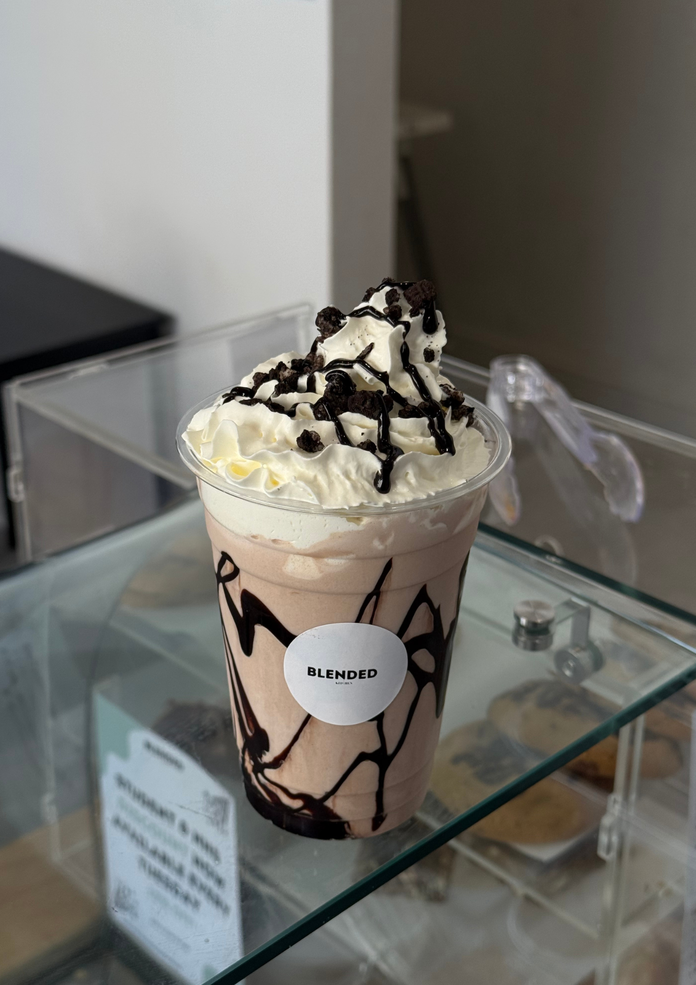Chocolate milkshake topped with whipped cream, chocolate syrup, and crumbles in a clear cup labeled 'Blended' on a glass display shelf.