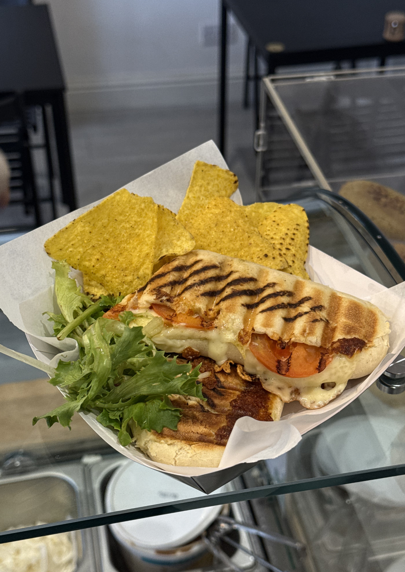A grilled chicken and tomato sandwich with melted cheese, accompanied by a side of lettuce, served with tortilla chips on a paper lining in a takeout container.