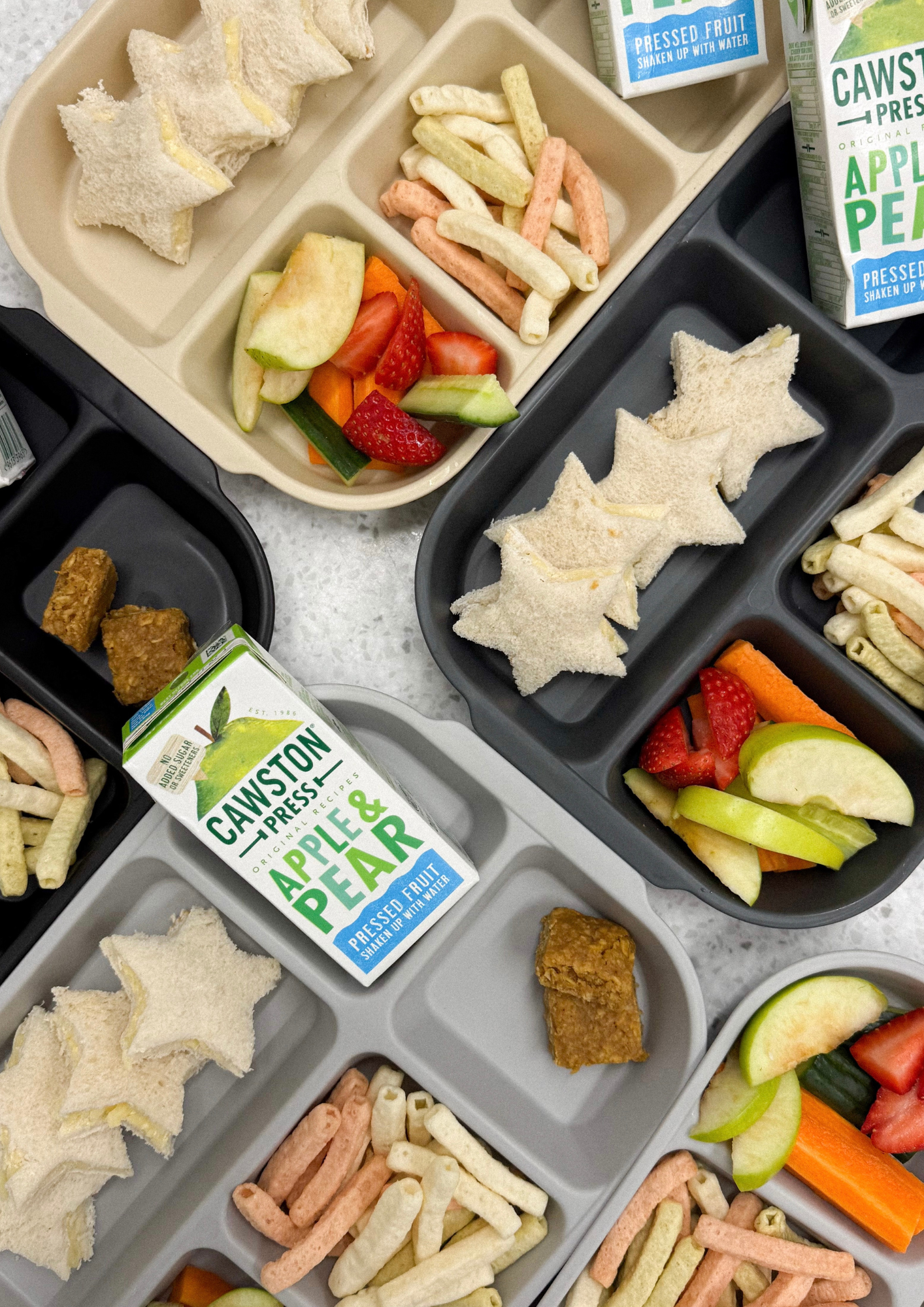 School lunch tray with sandwiches, fruit slices, veggie sticks, colorful cheese puffs, and cartons of apple and pear juice.