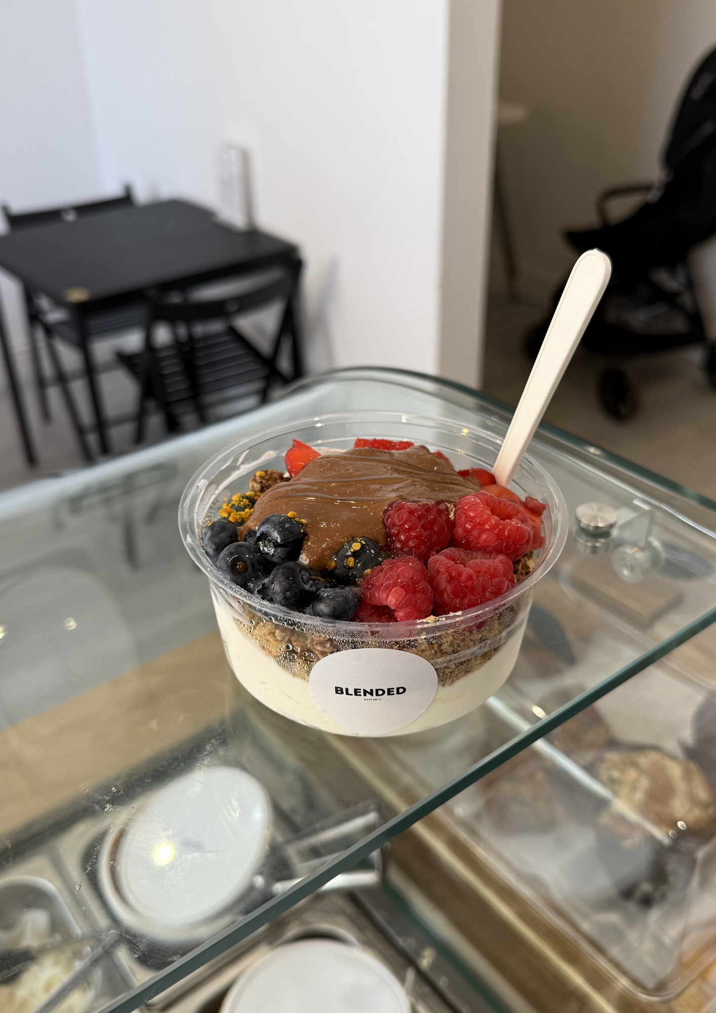 Close-up of a cup of layered dessert with fresh berries, chocolate, and crumb topping on a glass table with a blurred background of black chairs and a small table in a cafe or restaurant setting.