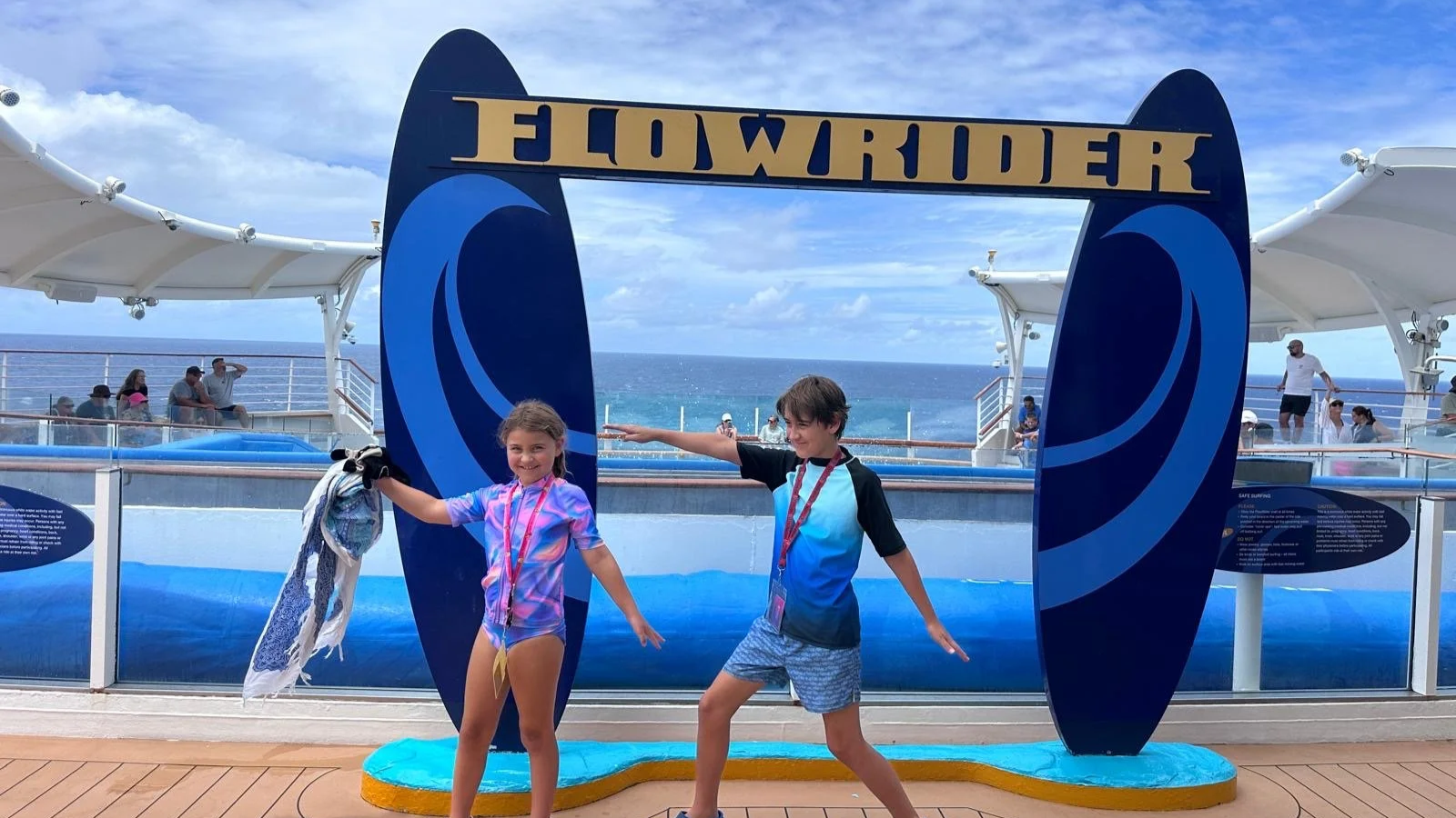 Two children pose playfully at a cruise ship's pool area, holding towels and wearing swimwear, under a blue and yellow sign that reads 'WONDER' and depicts waves, with the ocean and sky in the background.