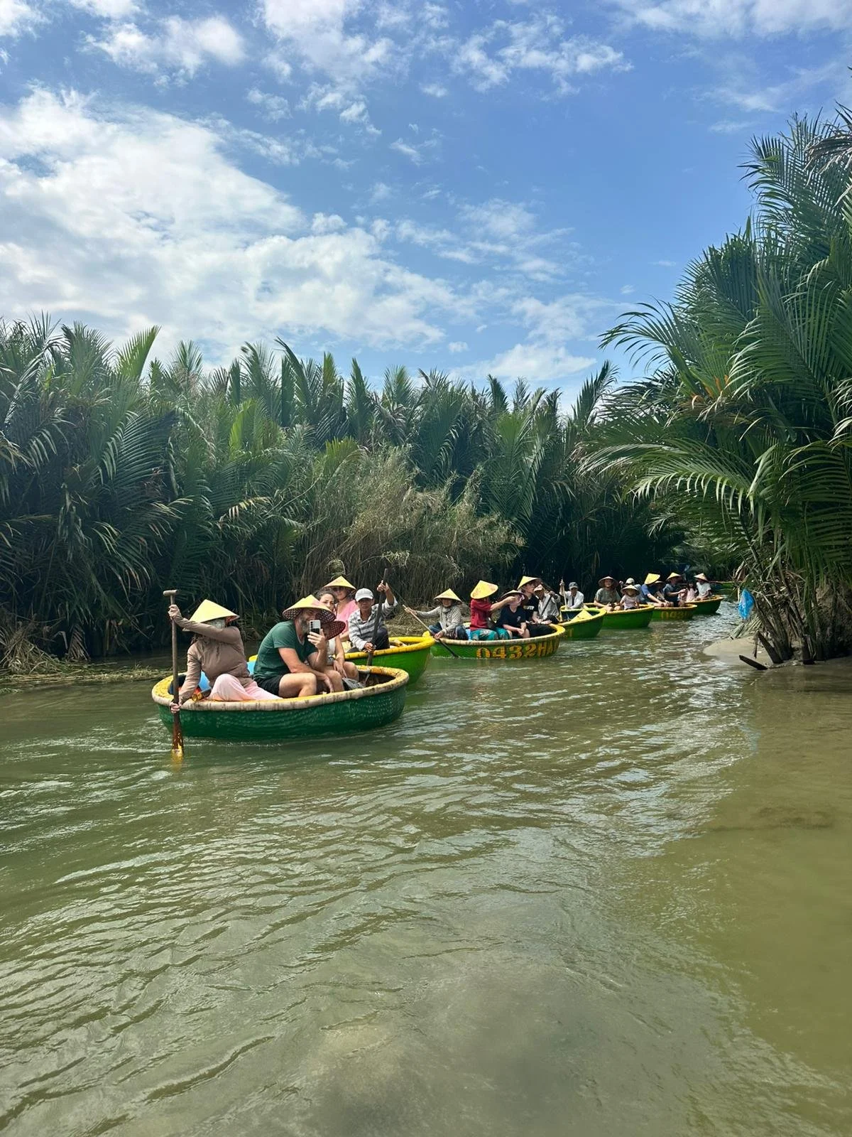 Tourists in round boats with straw hats riding through a narrow waterway surrounded by dense green tropical plants under a blue sky with clouds.