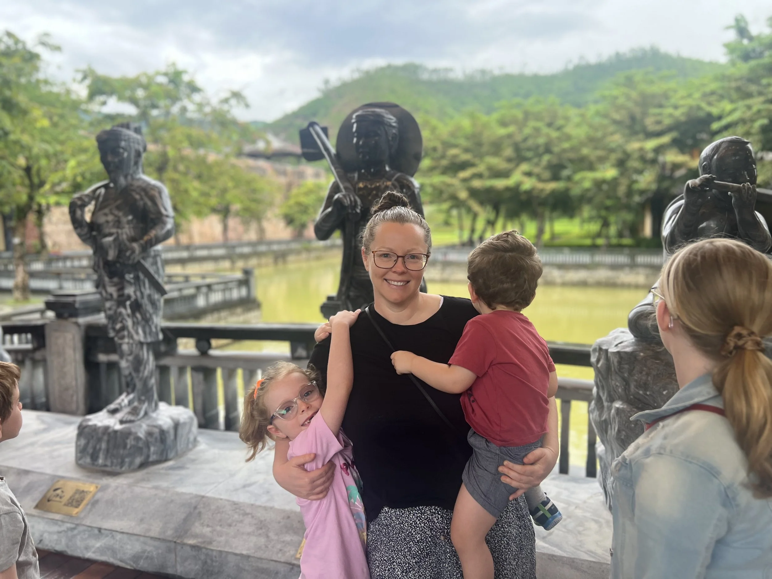 A woman holding a young girl in pink, with a young boy in red in her arms, surrounded by children and bronze statues of Native Americans in a park or historical site with green trees and hills in the background.
