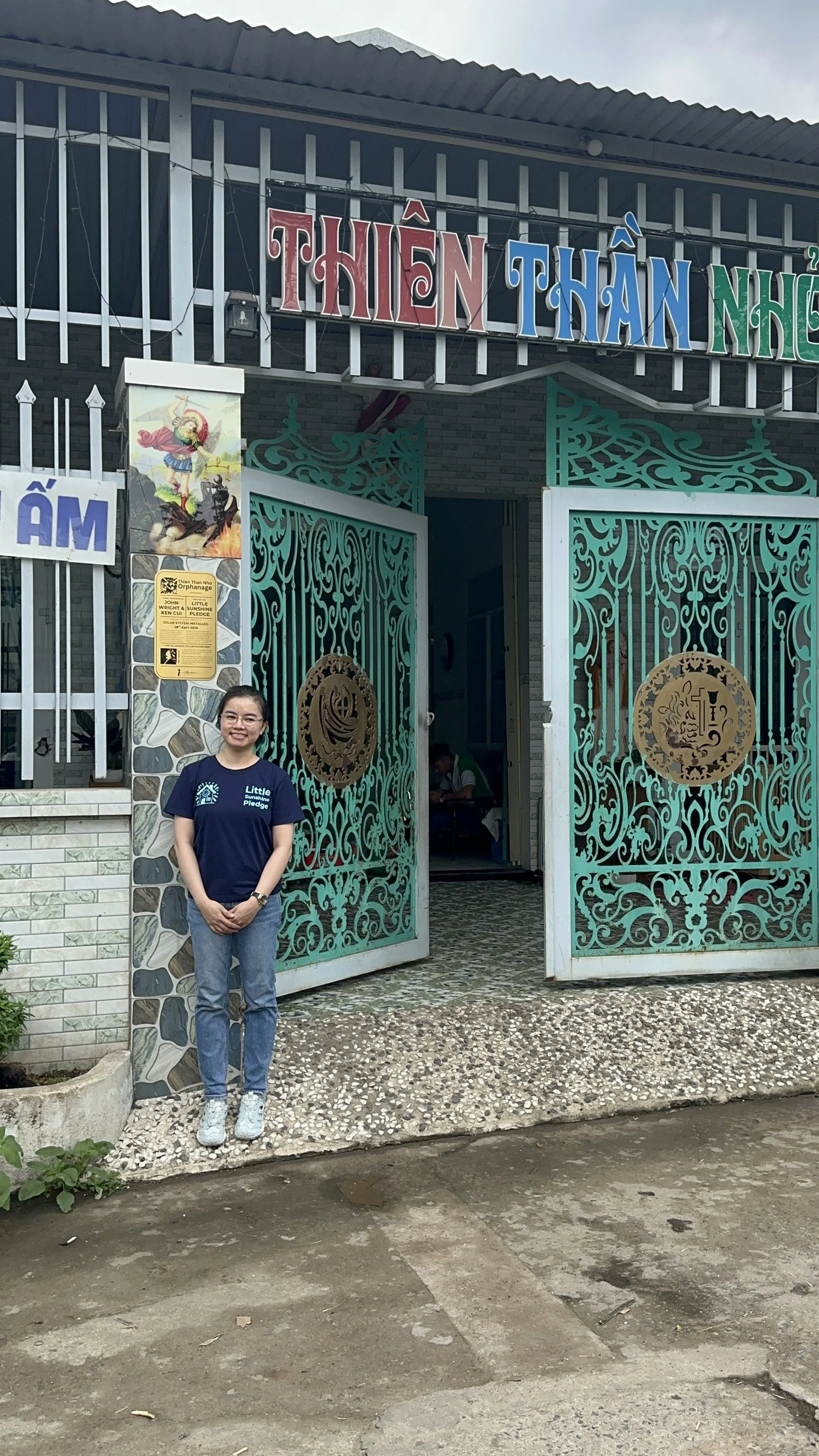 Phuong is standing at the front of Thien Than Nho with the plaque positioned behind her on the column of the orphanage