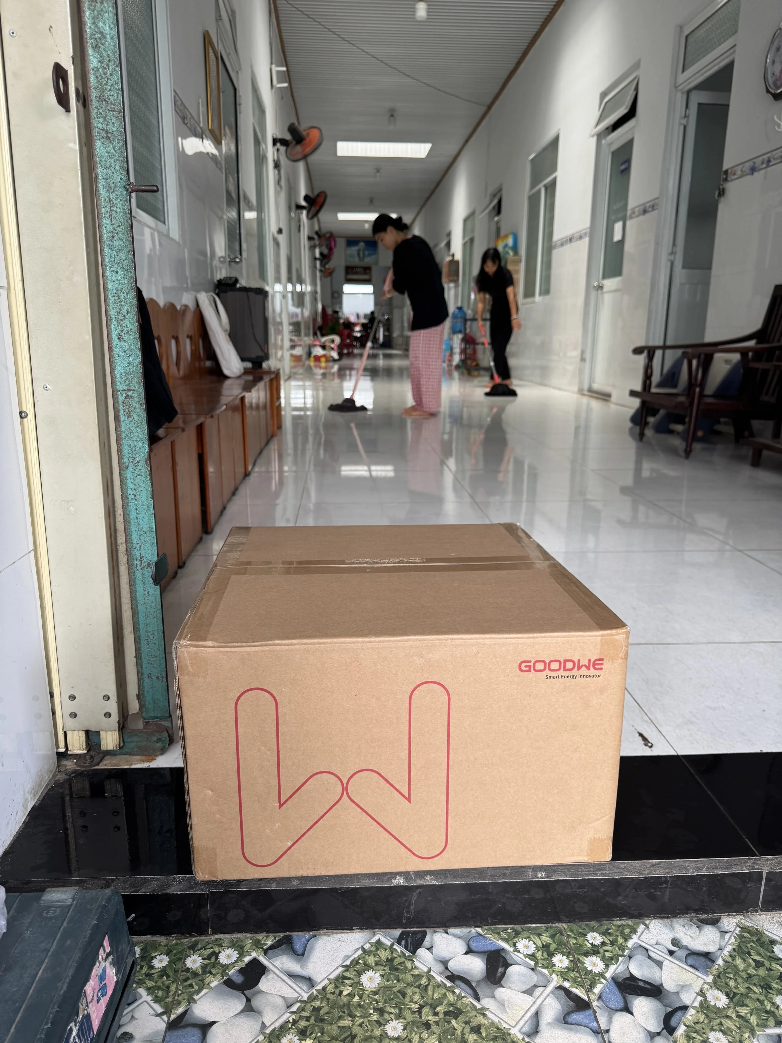 Photo of GoodWe inverter box at a doorstep with carers and kids cleaning the floor in the background