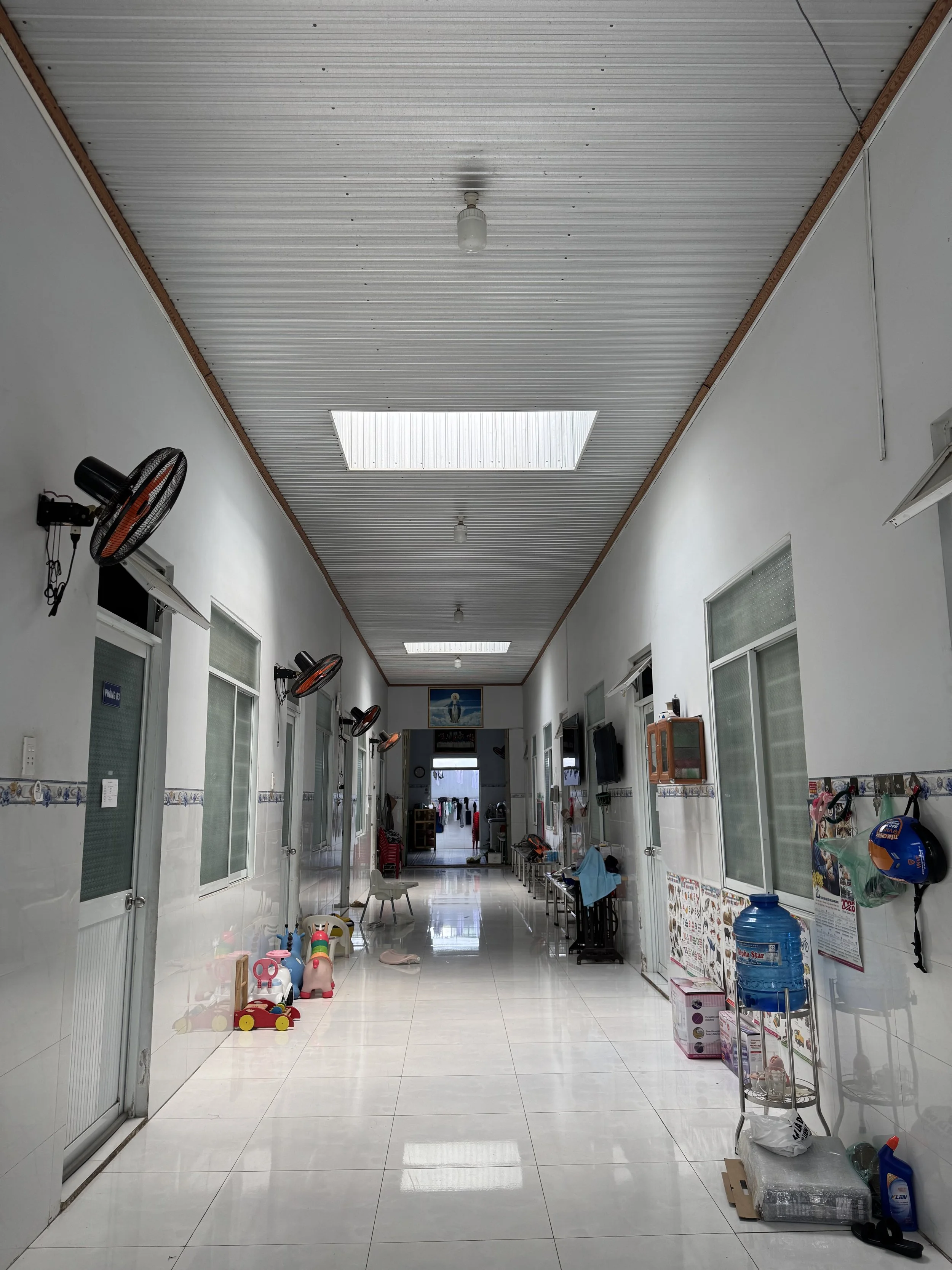 Photo shows an empty hallway of Thien Than Nho with fans on the walls and some toys lined up along the wall on the ground, there are skylights in the ceiling for natural lighting