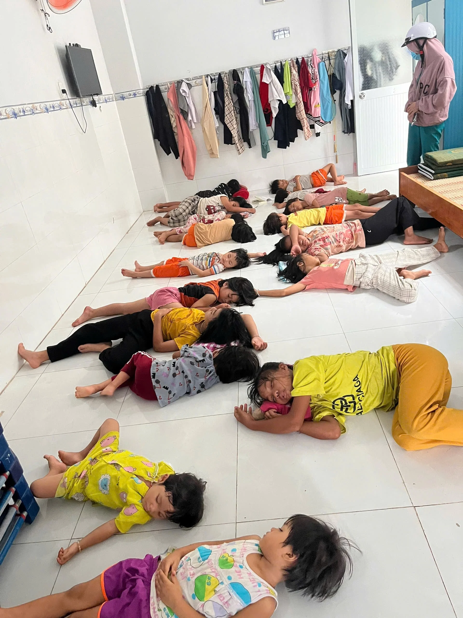 Showing a group of kids taking a nap on tile floor because of the heat with clothes hanging on the back wall